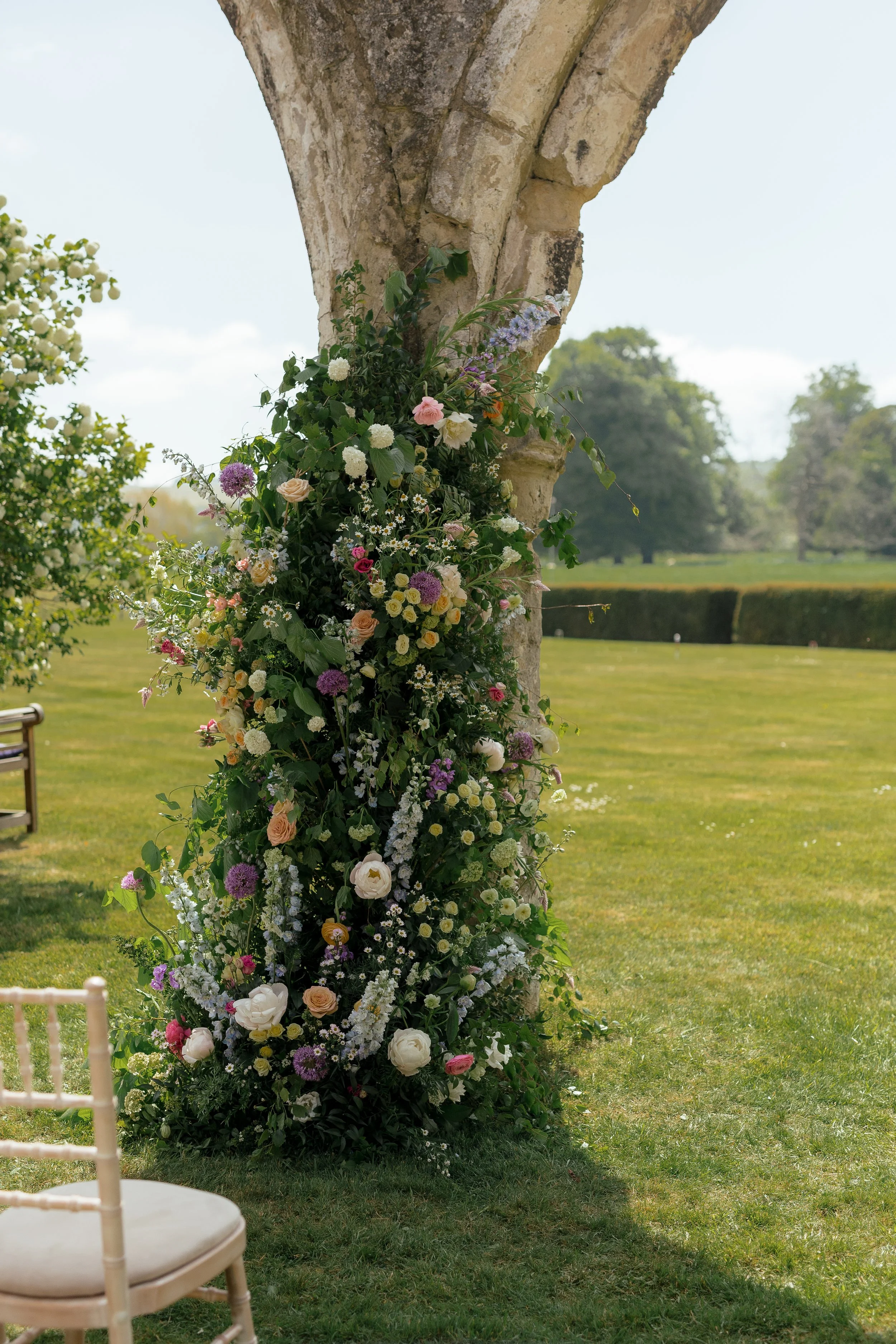 A vertical arrangement of various colorful flowers attached to the side of an old stone archway in an outdoor setting with a grassy field and trees in the background.