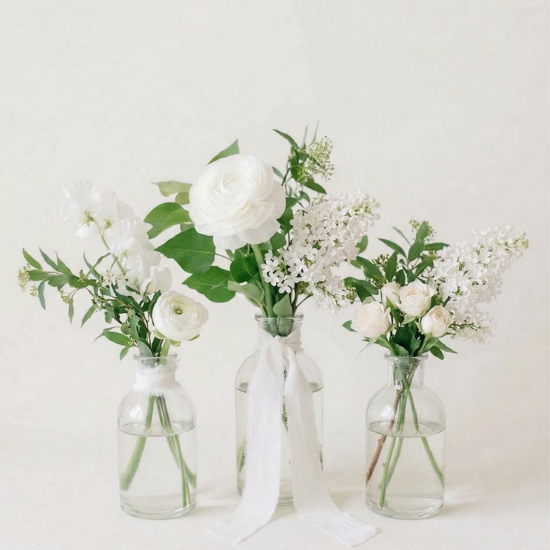 Three glass vases filled with white flowers and green leaves, against a plain background.