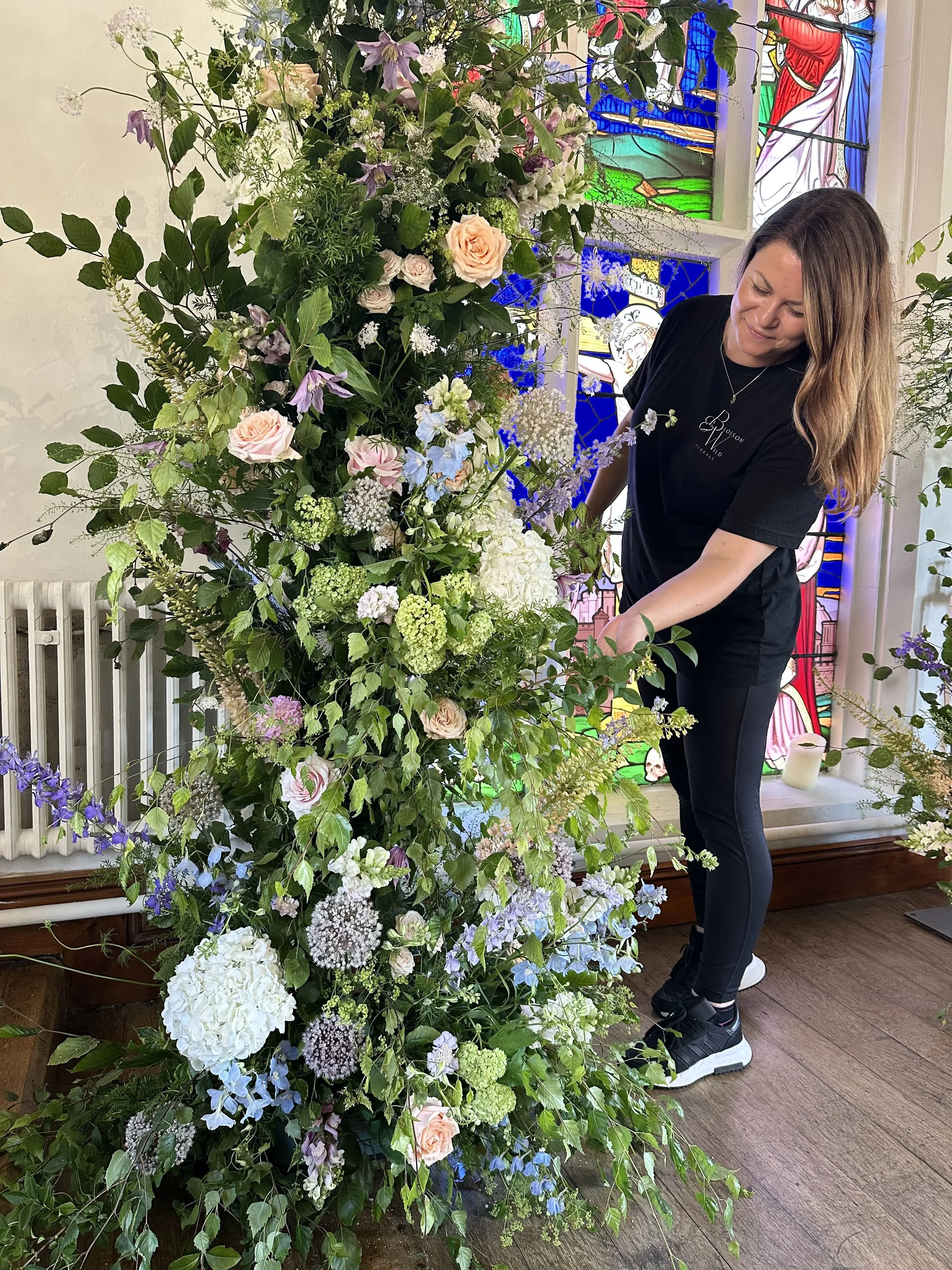A woman arranging a large, colorful flower display featuring roses, hydrangeas, and various greenery inside a room with stained glass windows.