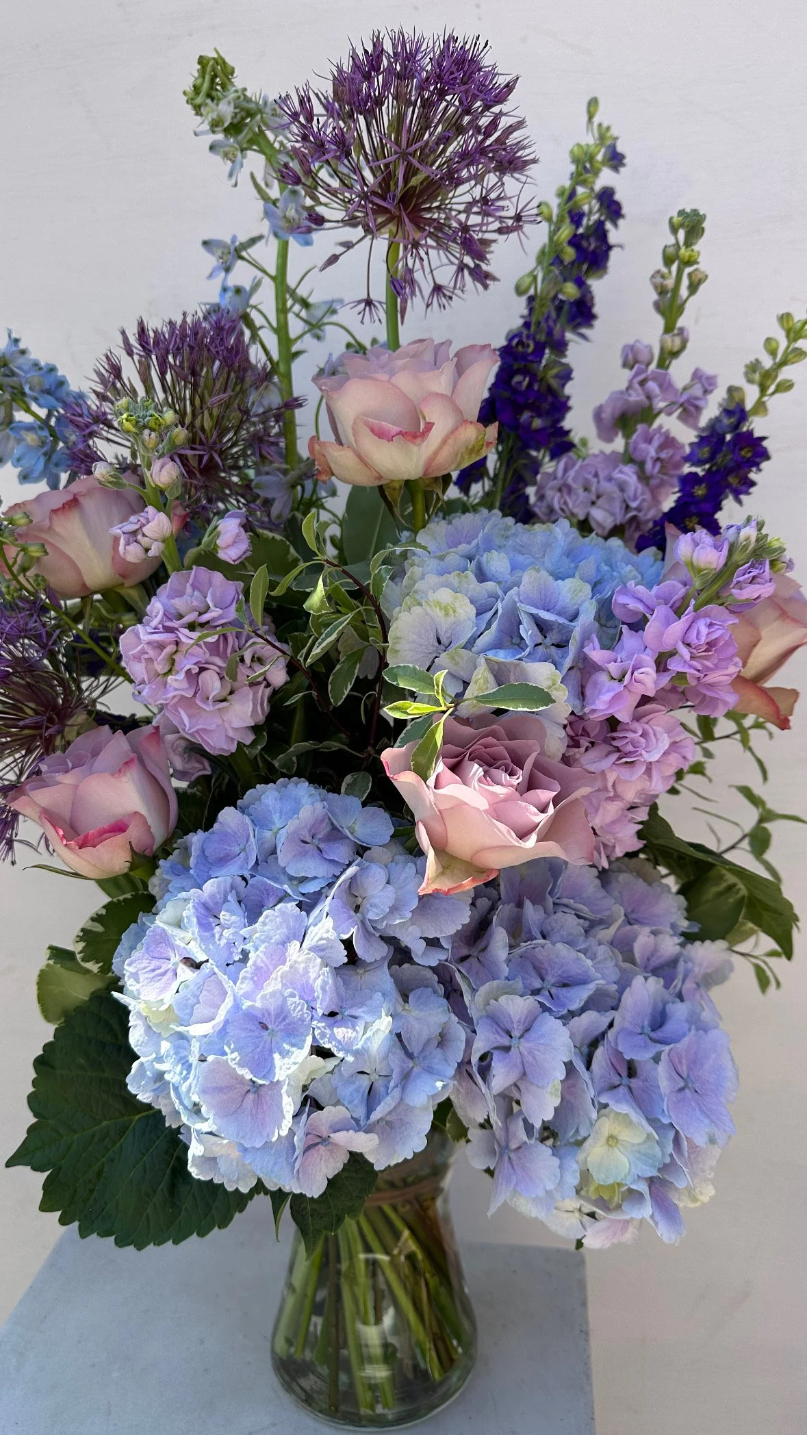 A glass vase filled with a bouquet of purple, pink, and blue flowers, including hydrangeas and alliums, against a plain white background.