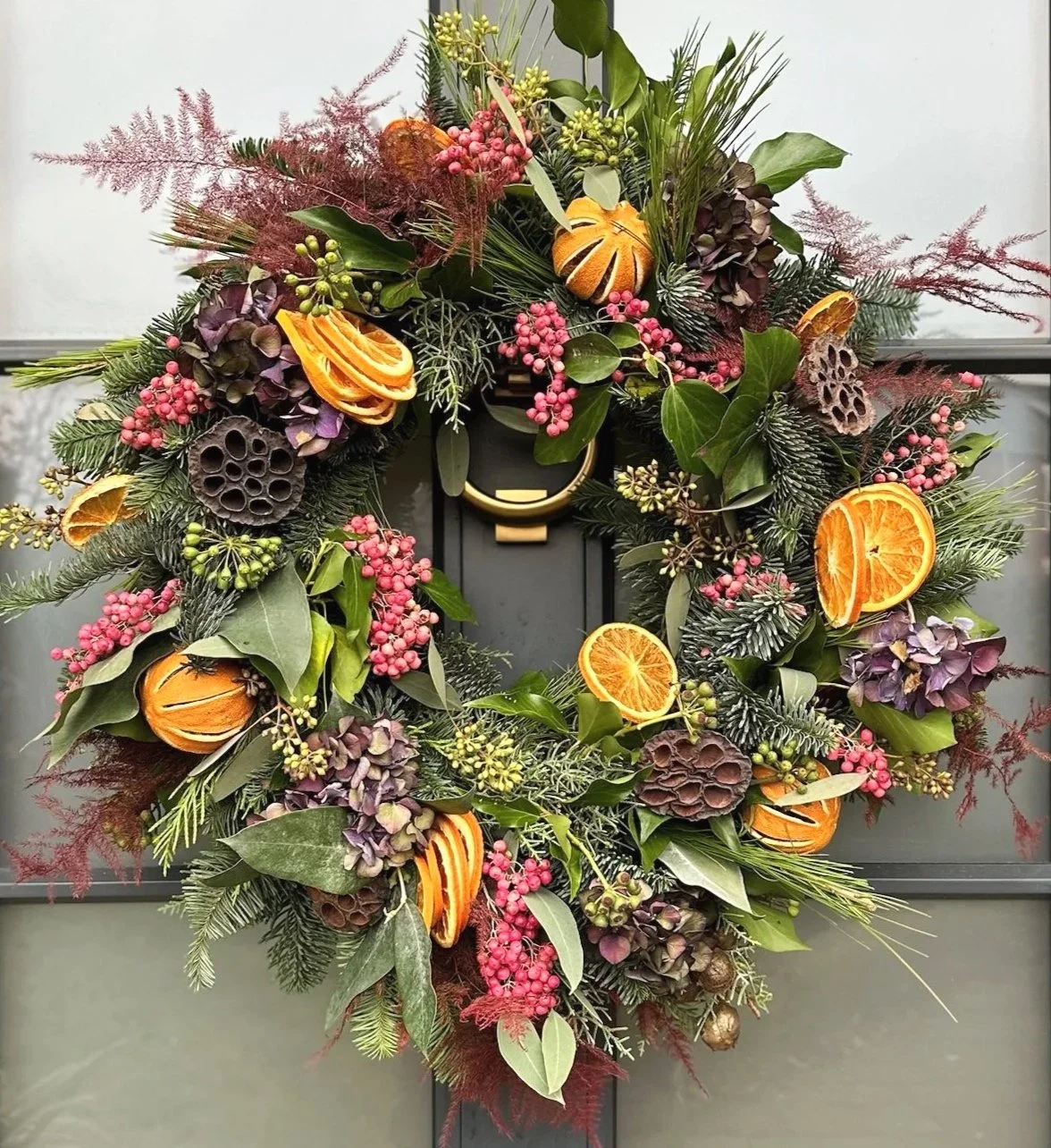 A festive holiday wreath decorated with dried orange slices, pink berries, dried flowers, pinecones, and various green foliage, hanging on a dark door.