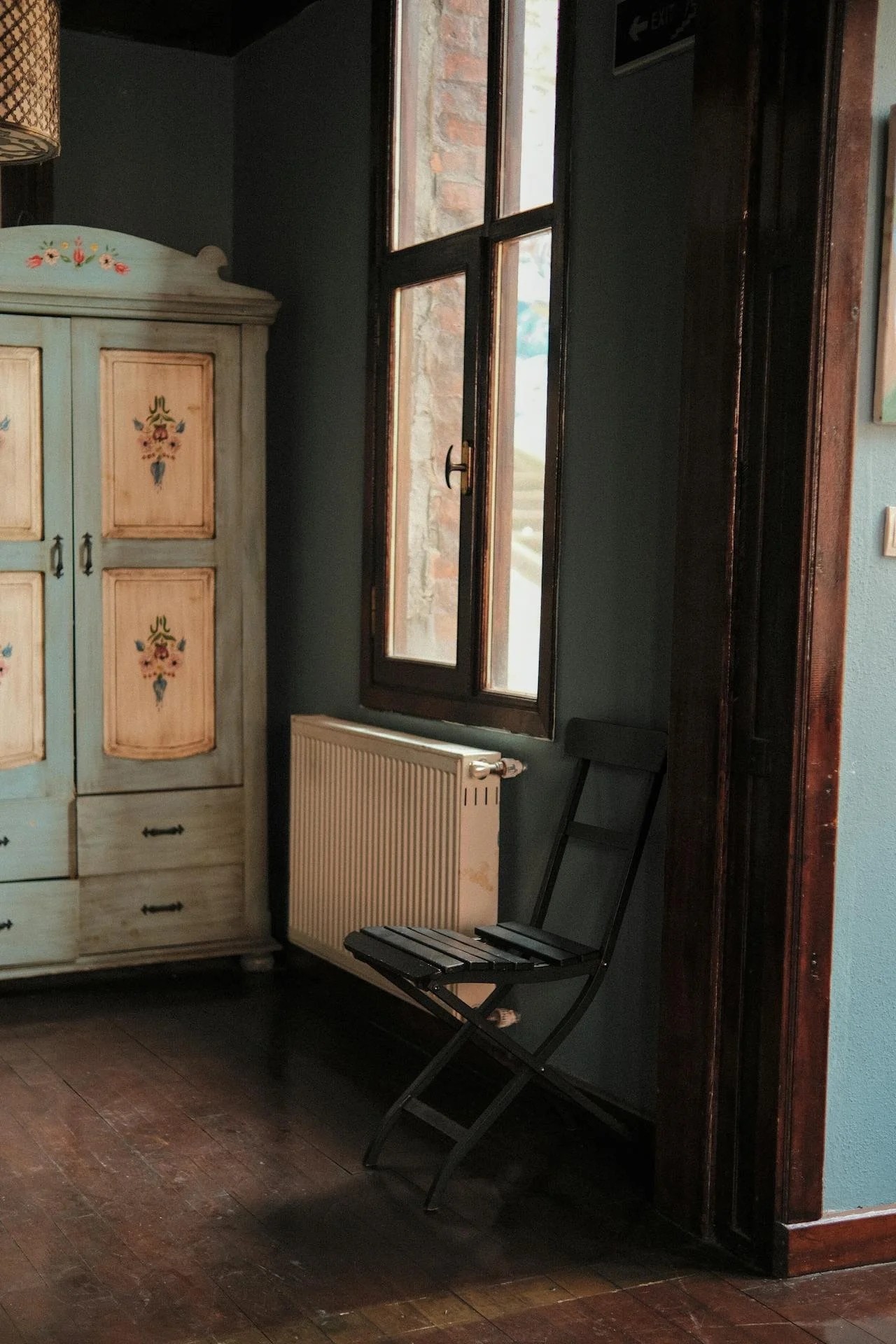 A corner of a room with a vintage painted cabinet, a radiator, a window with open glass panes, and a black folding chair.