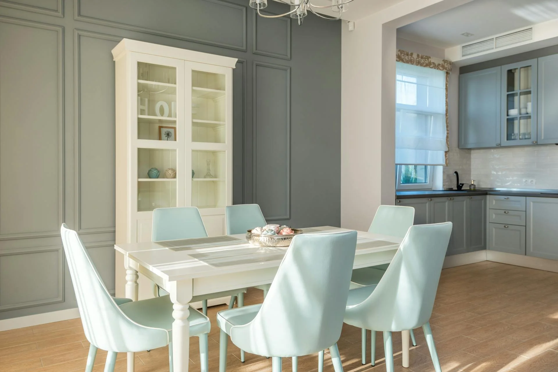 A bright dining room with a white table surrounded by six light blue cushioned chairs, a white china cabinet, and a kitchen in the background with blue cabinets and a window.