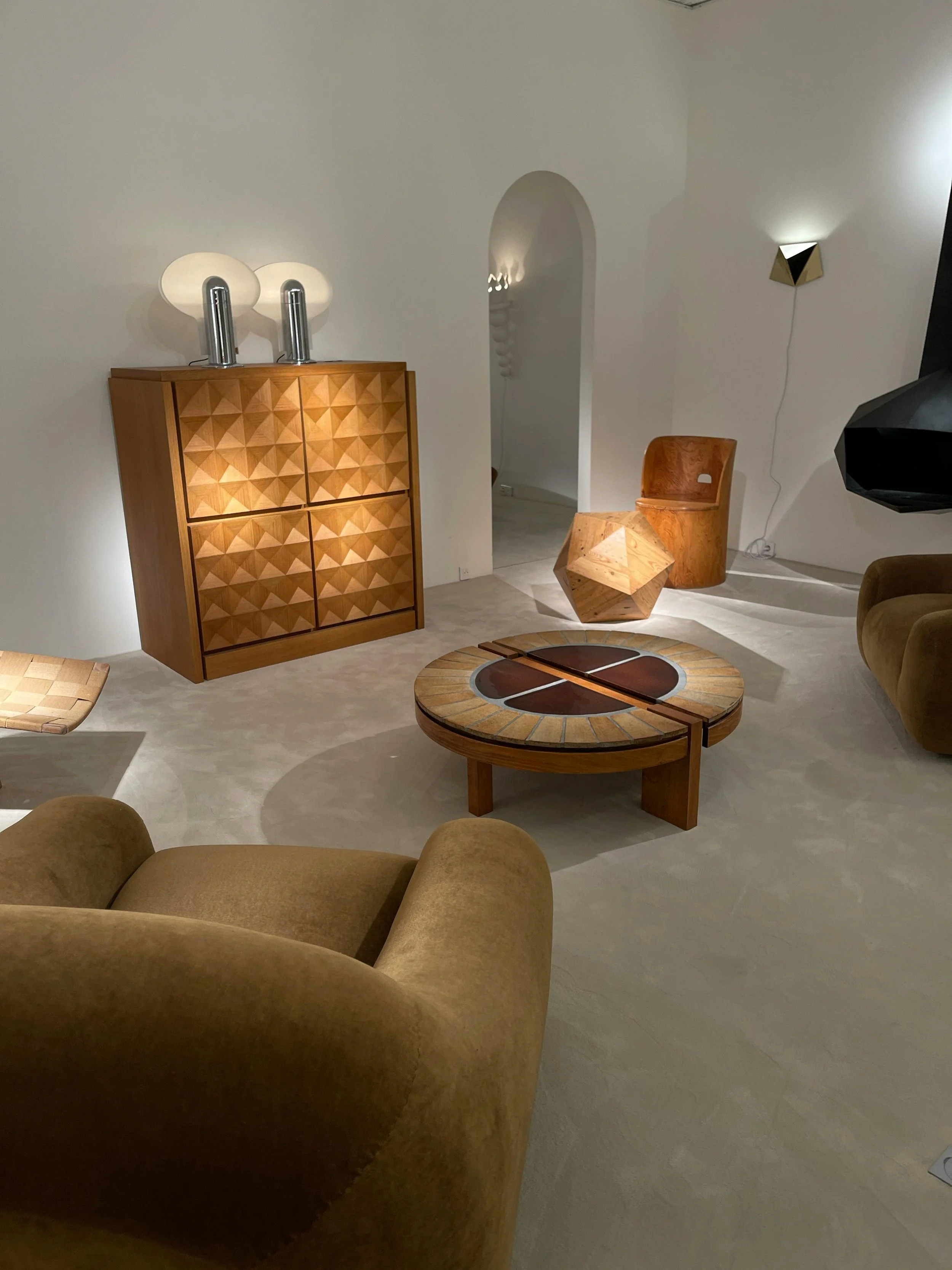 Living room with mid-century modern furniture, including a round wooden coffee table, a tan sofa, a wooden armchair, a modular geometric side table, and a wooden cabinet with two lamps on top.
