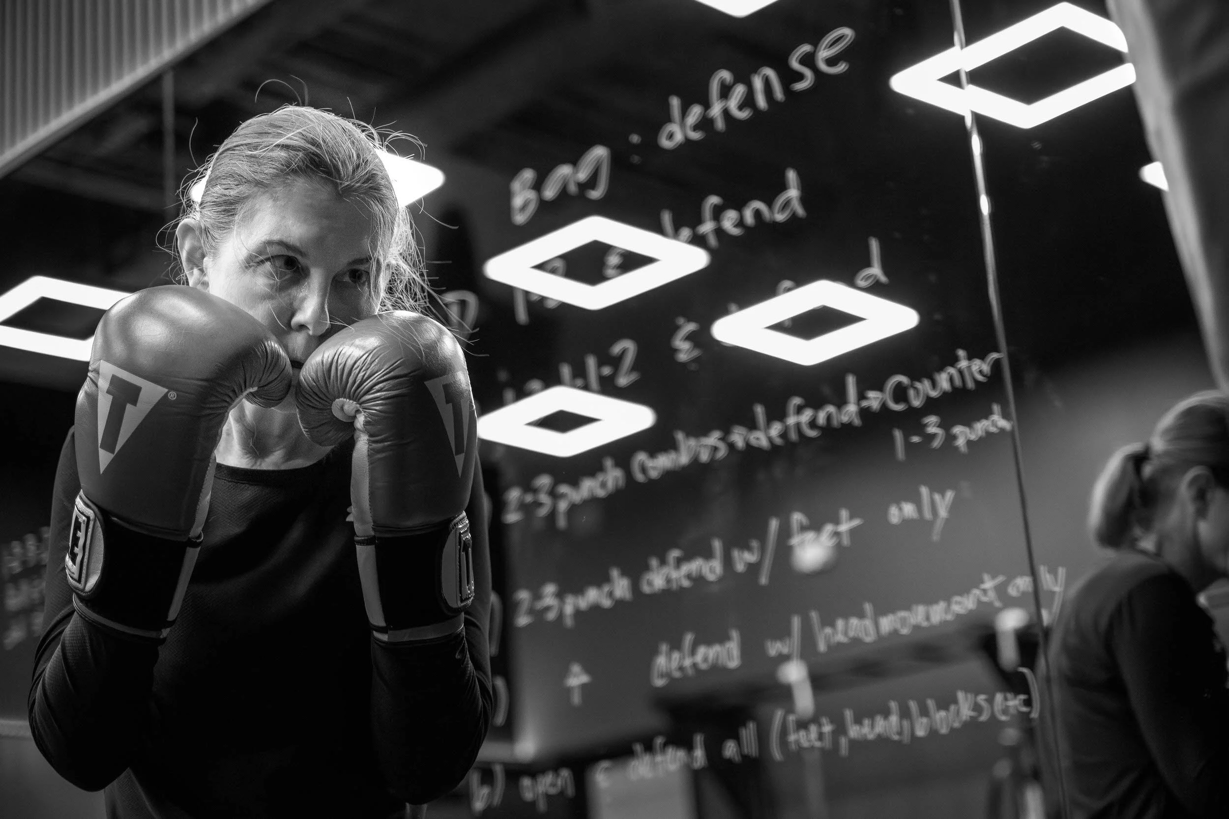 A woman holding punching mitts is training with a man in a gym, with punching bags in the background.