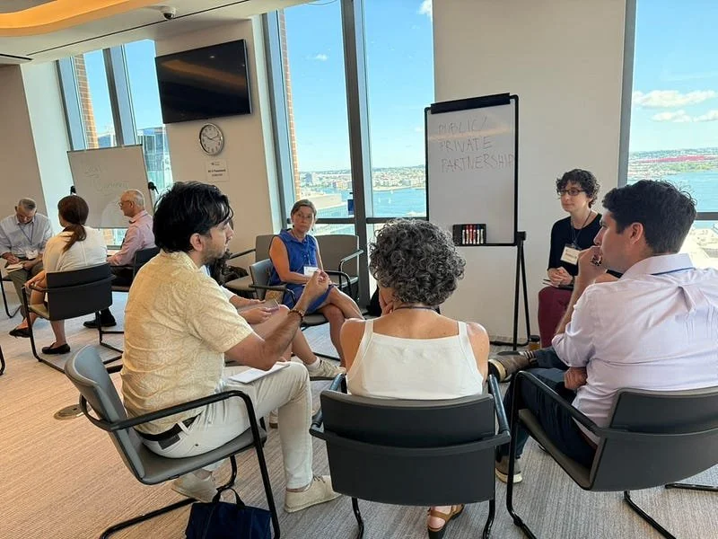 At InnSure's Insurability Planning workshop in Boston (2025), a group of six people in a circle discuss "Public Private Partnerships" while overlooking the harbor. Other groups can be seen in the background.