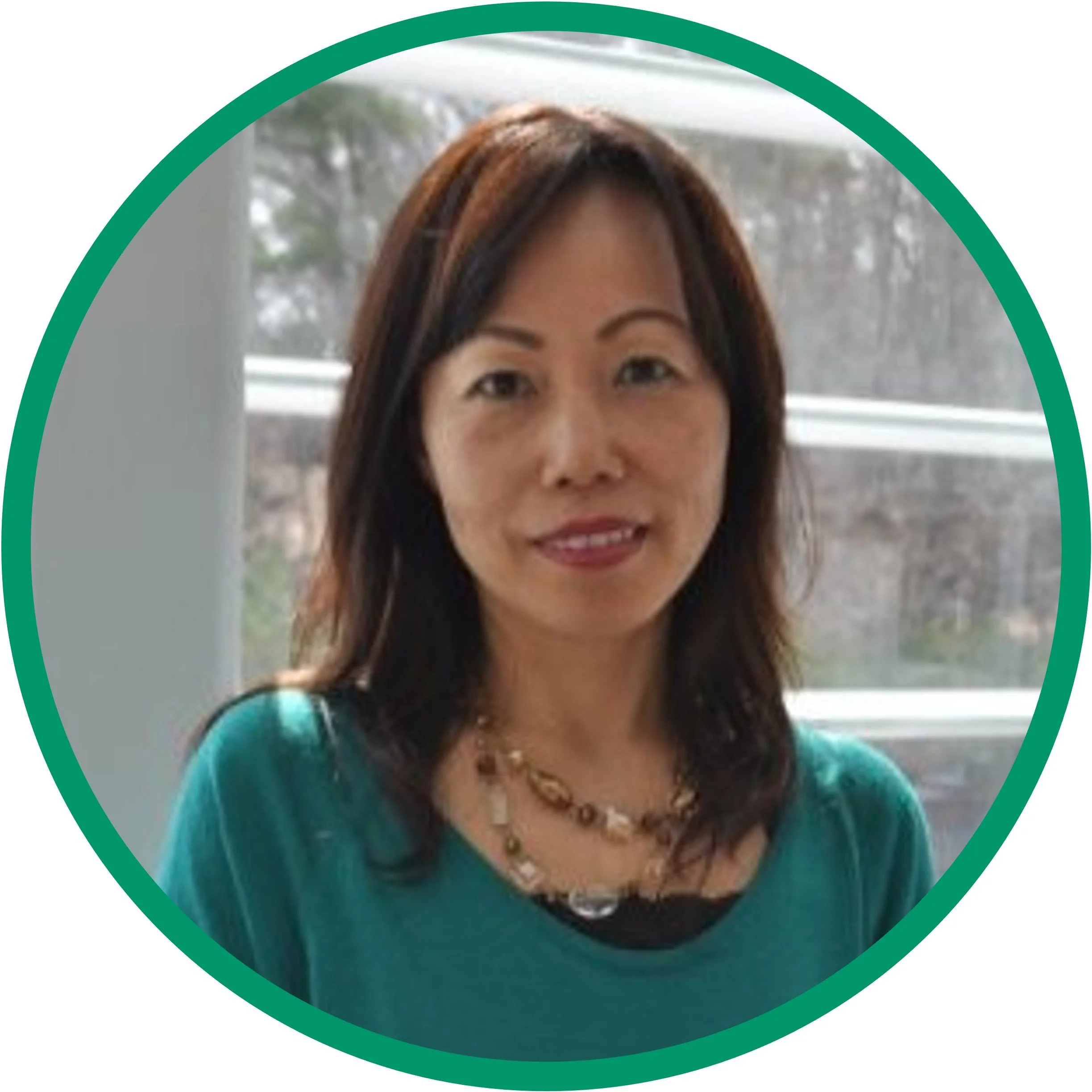 A woman with shoulder-length dark hair wearing a teal top and a beaded necklace standing in front of a window.