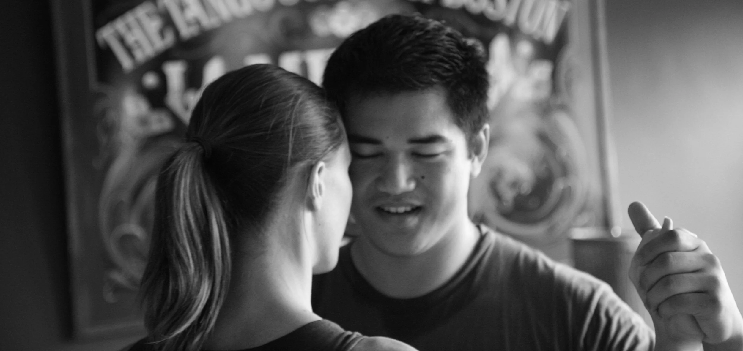 Black and white image of two people dancing closely, one with a ponytail and the other with short hair, smiling and holding hands.