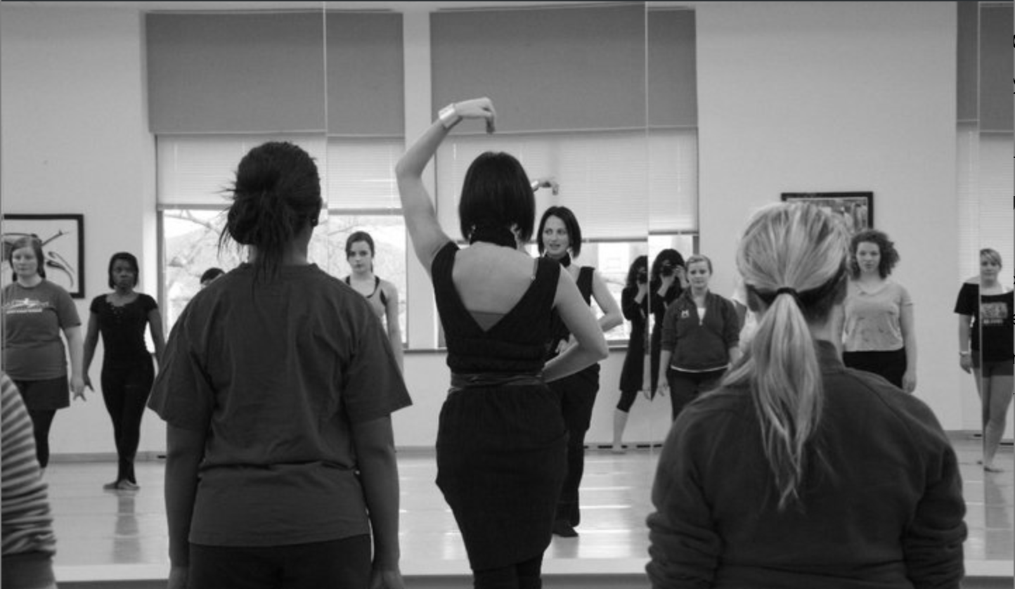 A dance instructor leads a group of students in a dance class, with participants facing a mirrored wall.