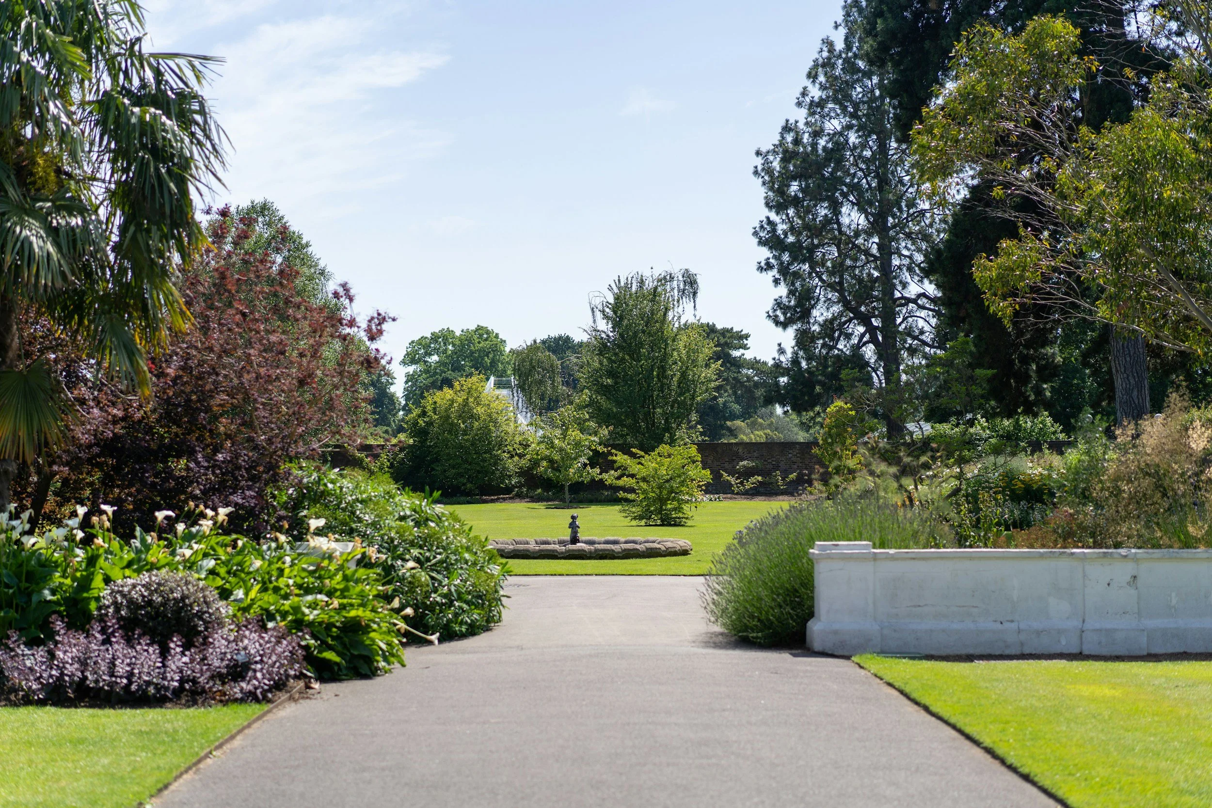 A pathway in a lush garden with green grass, trees, bushes, and a small fountain in the distance on a sunny day.