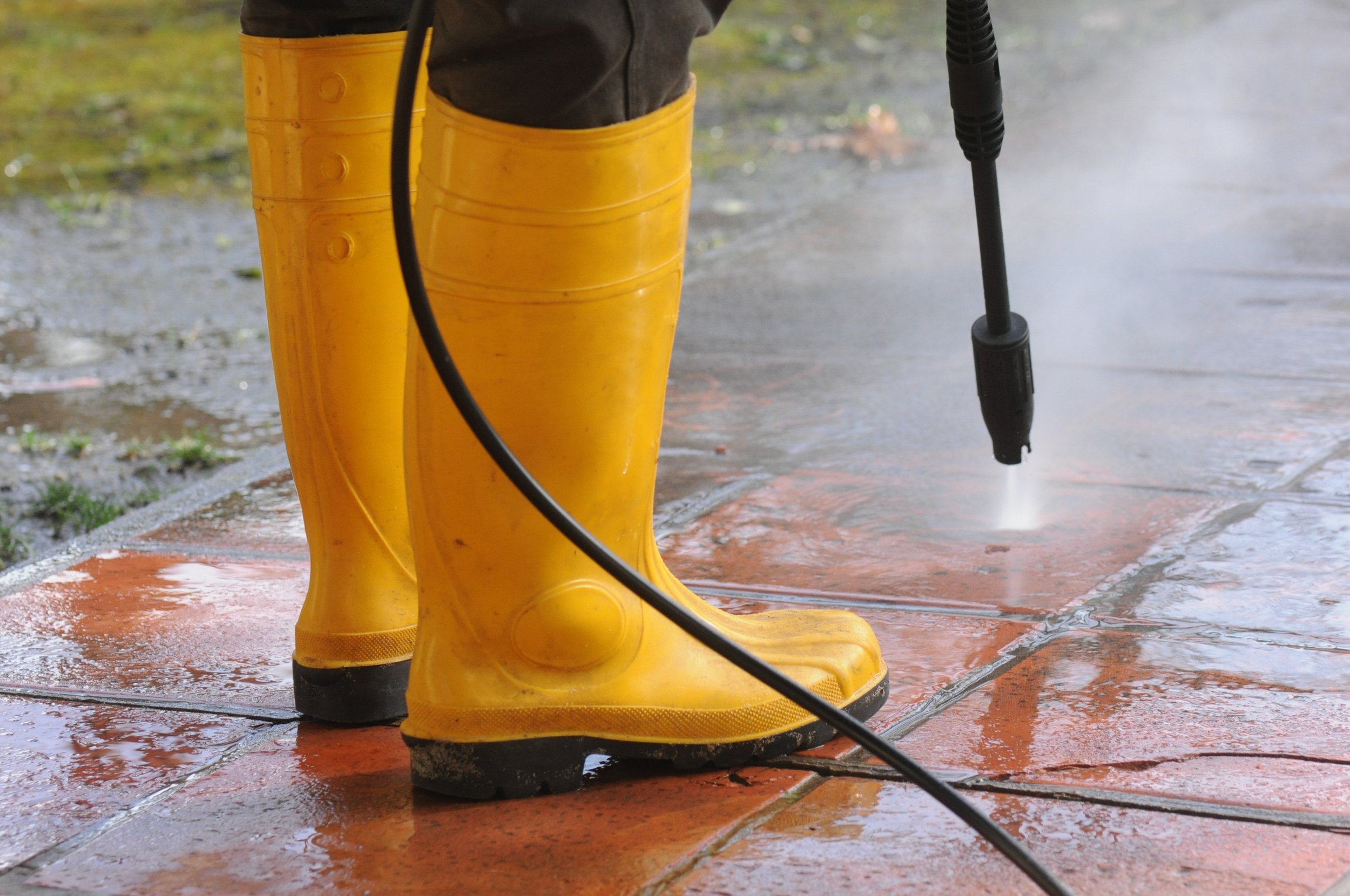 Person wearing yellow rubber boots using a pressure washer on wet patio driveway.