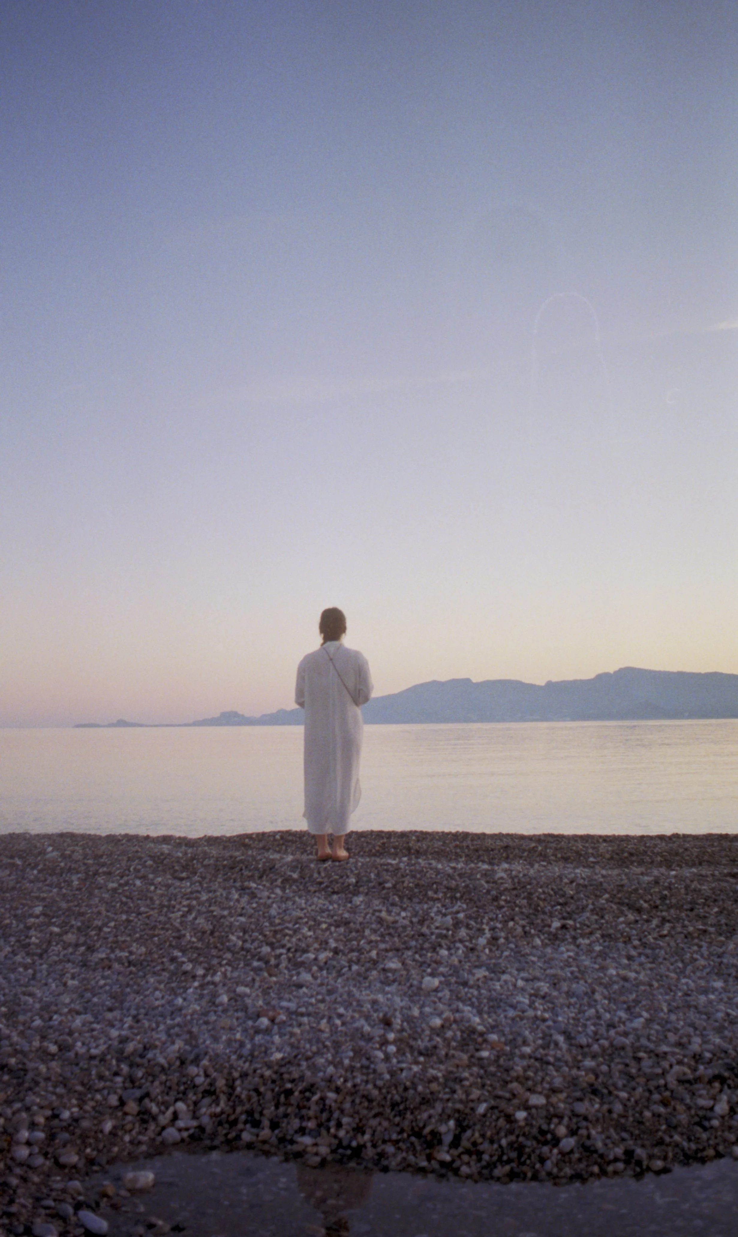 Eine Frau in weißem, fließenden Kleid steht zur blauen Stunde am Ufer des Meeres, blickt auf das Wasser und die Berge im Hintergrund.