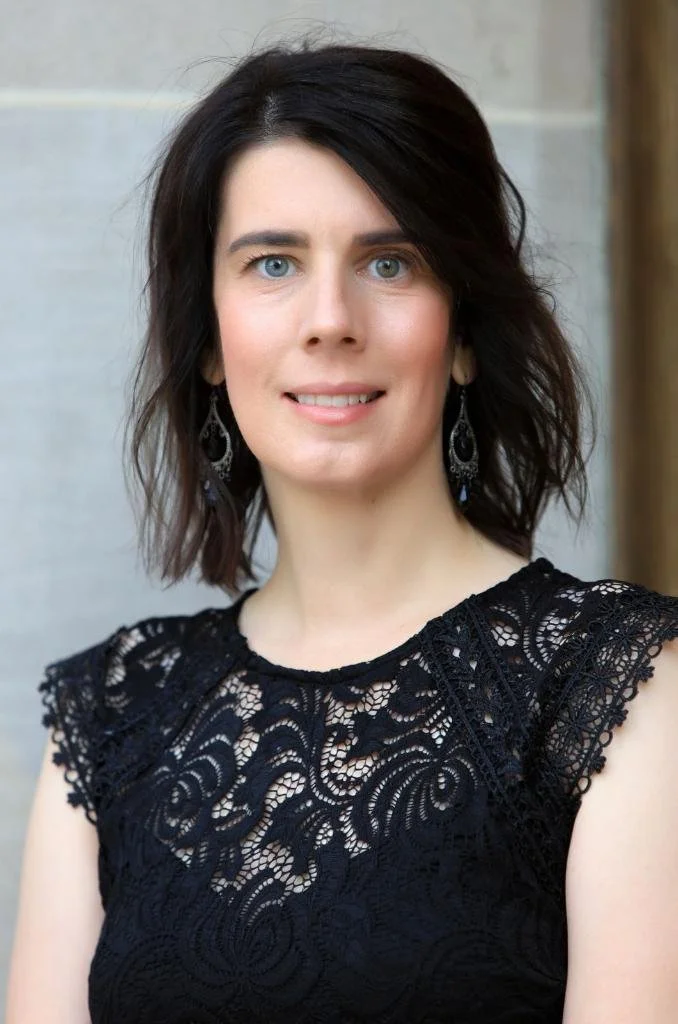 A woman with dark hair wearing a black lace top and dangling earrings, smiling outdoors.