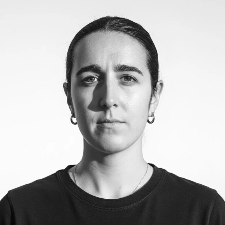 Black and white portrait of a young woman with dark hair tied back, wearing hoop earrings and a black shirt, looking directly at the camera with a neutral expression.