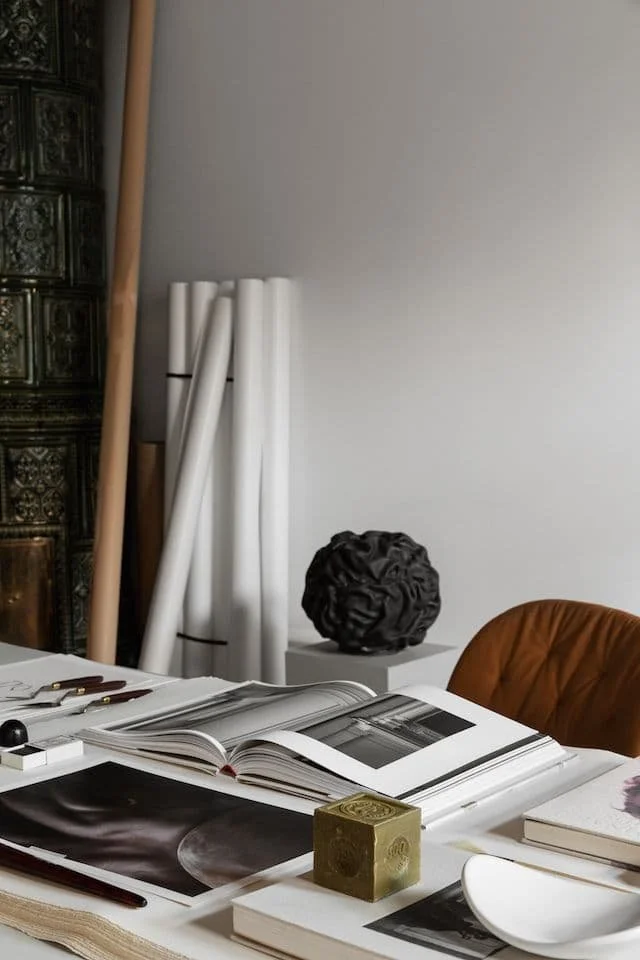 A workspace with an open photo book, a closed book, a gold decorative object, and various art supplies on a table. In the background, there are rolled white papers and a black decorative sculpture, with a brown chair nearby.