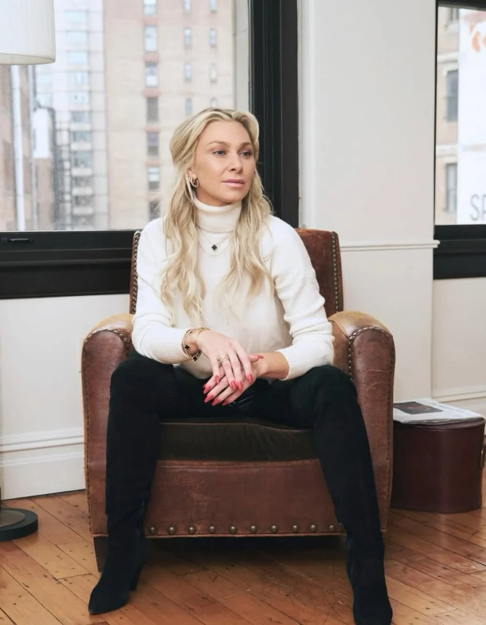 A woman with blonde hair sitting in a leather armchair near a window in an urban apartment, wearing a white turtleneck sweater and black pants.