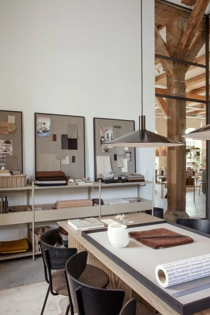 Interior of a store or studio with a table, black chairs, and shelves displaying fabric samples and artwork, with wooden beams and a large window.