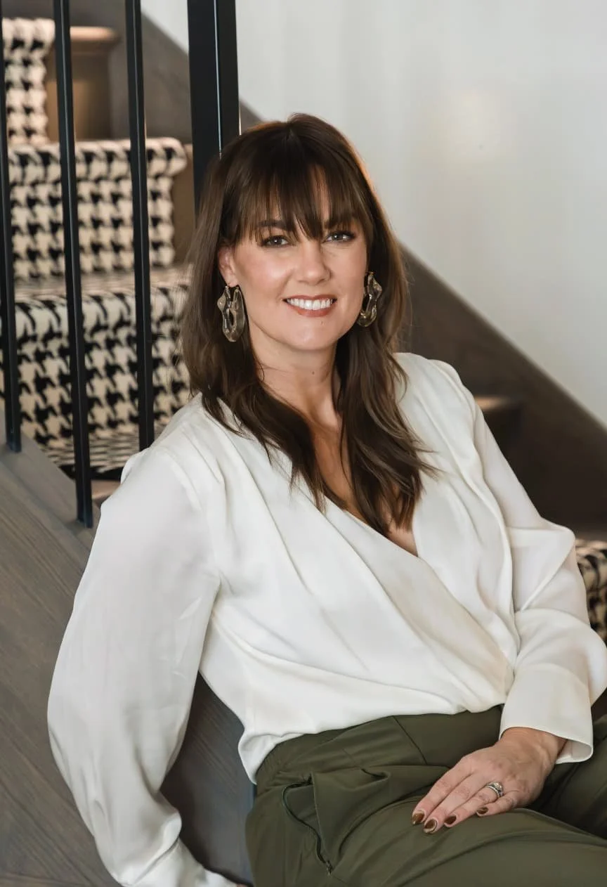 A woman with brown hair and bangs sitting on a bench indoors, smiling at the camera, wearing a white blouse and olive green pants, with earrings and a ring, near a staircase with black railings and patterned steps.