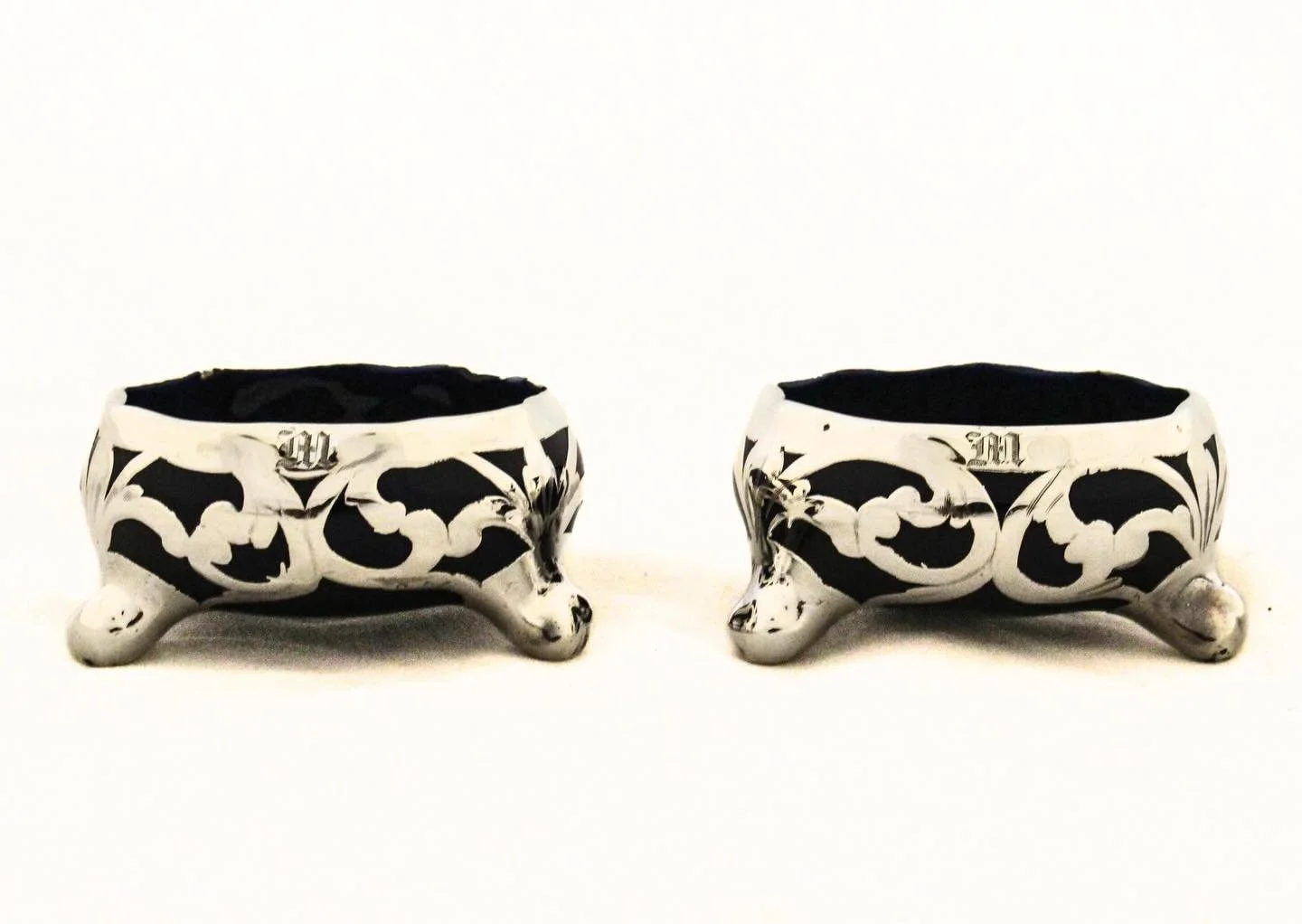 A refined pair of sterling silver overlay salt cellars, each of circular form with deep cobalt blue glass bodies permanently fused with intricately designed sterling silver overlay. The cellars rest on three applied silver feet, giving them a gracefu