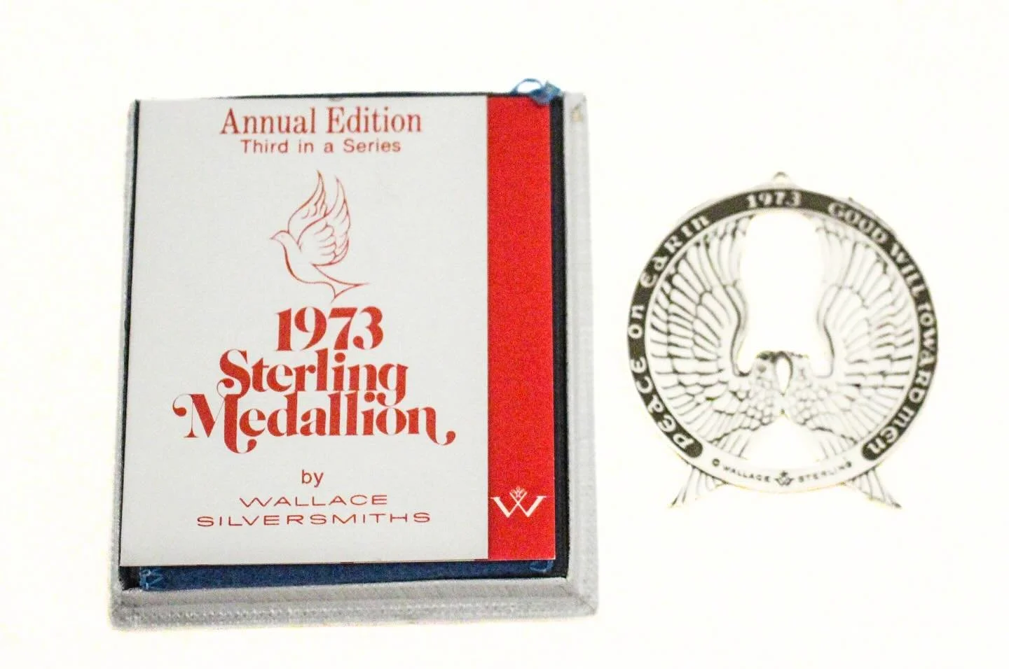 This is the 1973 Sterling Silver Christmas Medallion by Wallace Silversmiths, the third in their annual series. The ornament features two beautifully detailed, symmetrical angel wings forming a circular design, surrounded by the inscription &ldquo;Pe