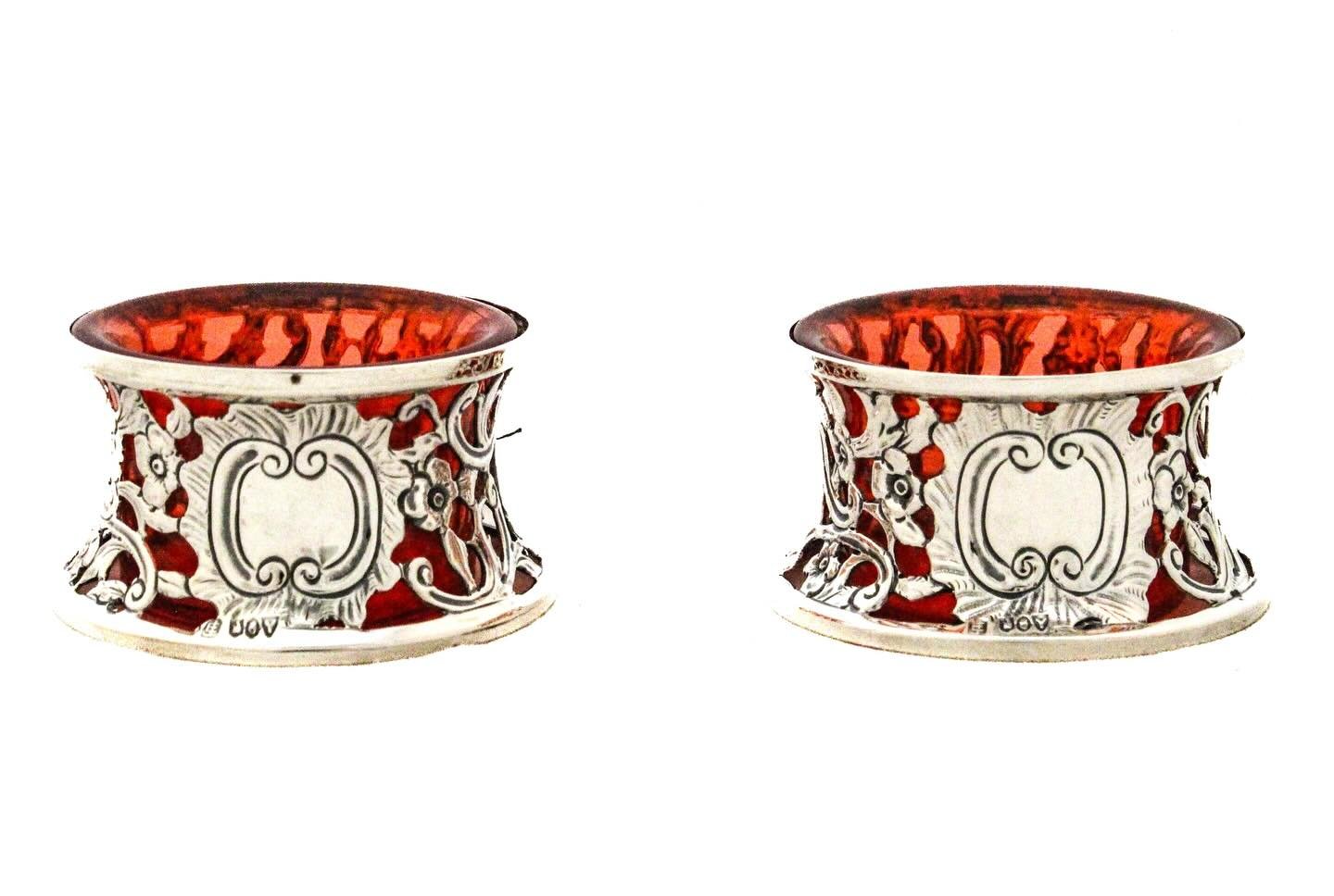A refined pair of antique American sterling silver salt cellars, decorated with elegant repouss&eacute; scrolling foliate motifs and classical blank cartouches. Each retains its original cranberry red glass liner, the rich ruby tone providing a strik