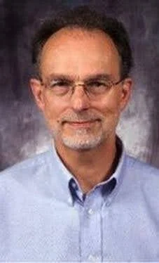 A middle-aged man with glasses and a beard wearing a light blue shirt against a dark background.