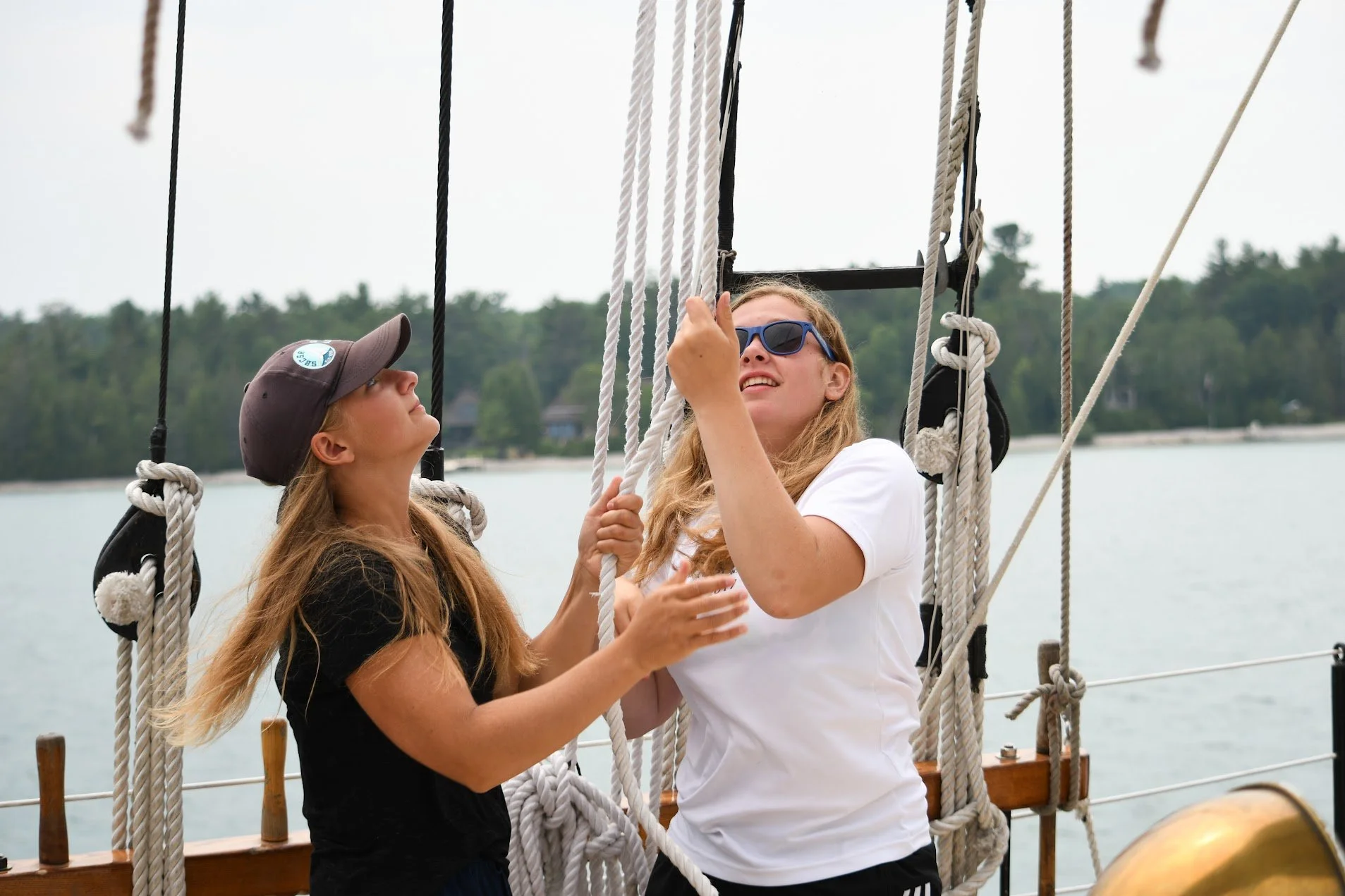 Two people working with ropes on a sailing ship, one wearing a cap and the other sunglasses, with water and trees in the background.
