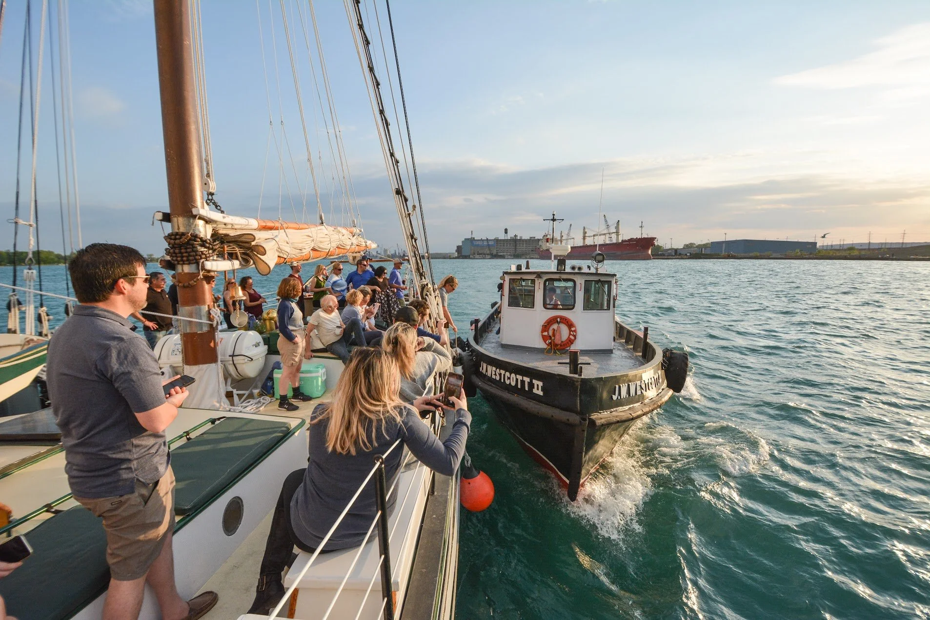 Group of people on a sailboat interacting with a smaller boat on a body of water during sunset, with industrial buildings and ships in the background.