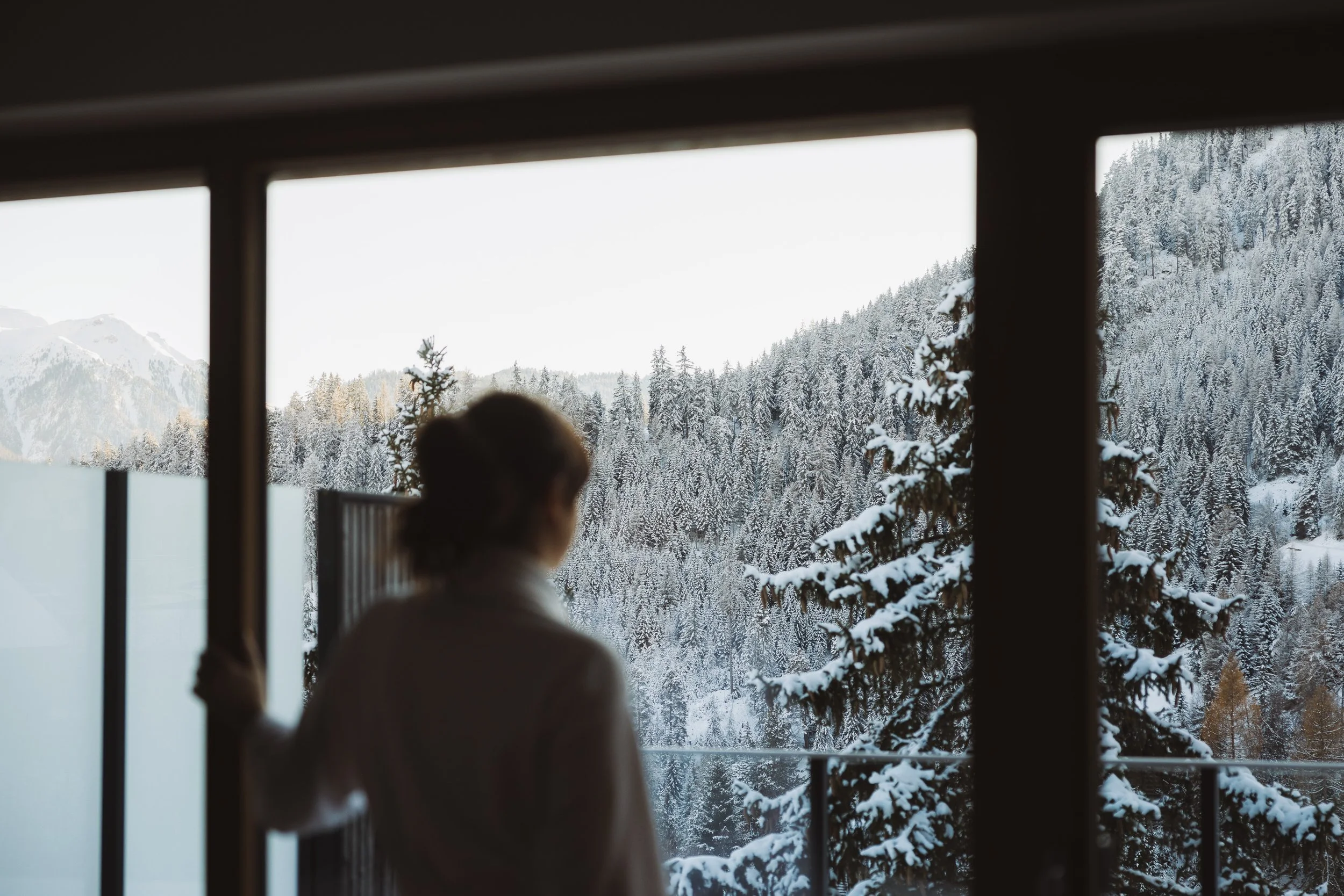 Frau blickt aus einem Fenster auf eine verschneite Winterlandschaft mit Tannenbäumen und Bergen.