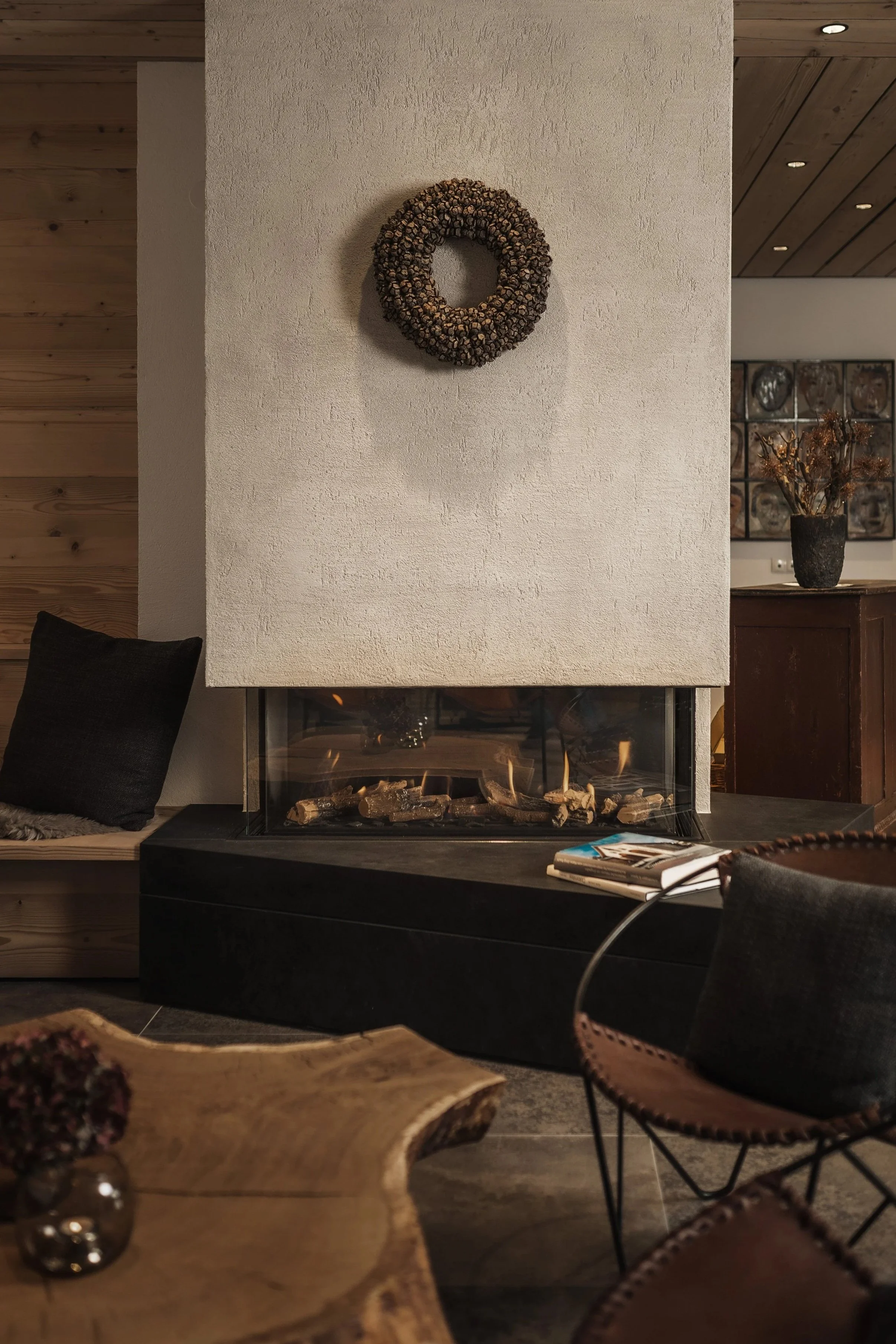 Gemütliches Wohnzimmer mit Kamin, Holzdekor, schwarzem Kissen auf einer Bank, Buch und Zeitschriften auf dem Couchtisch, Wanddekoration und Vase mit getrockneten Zweigen im Hintergrund.