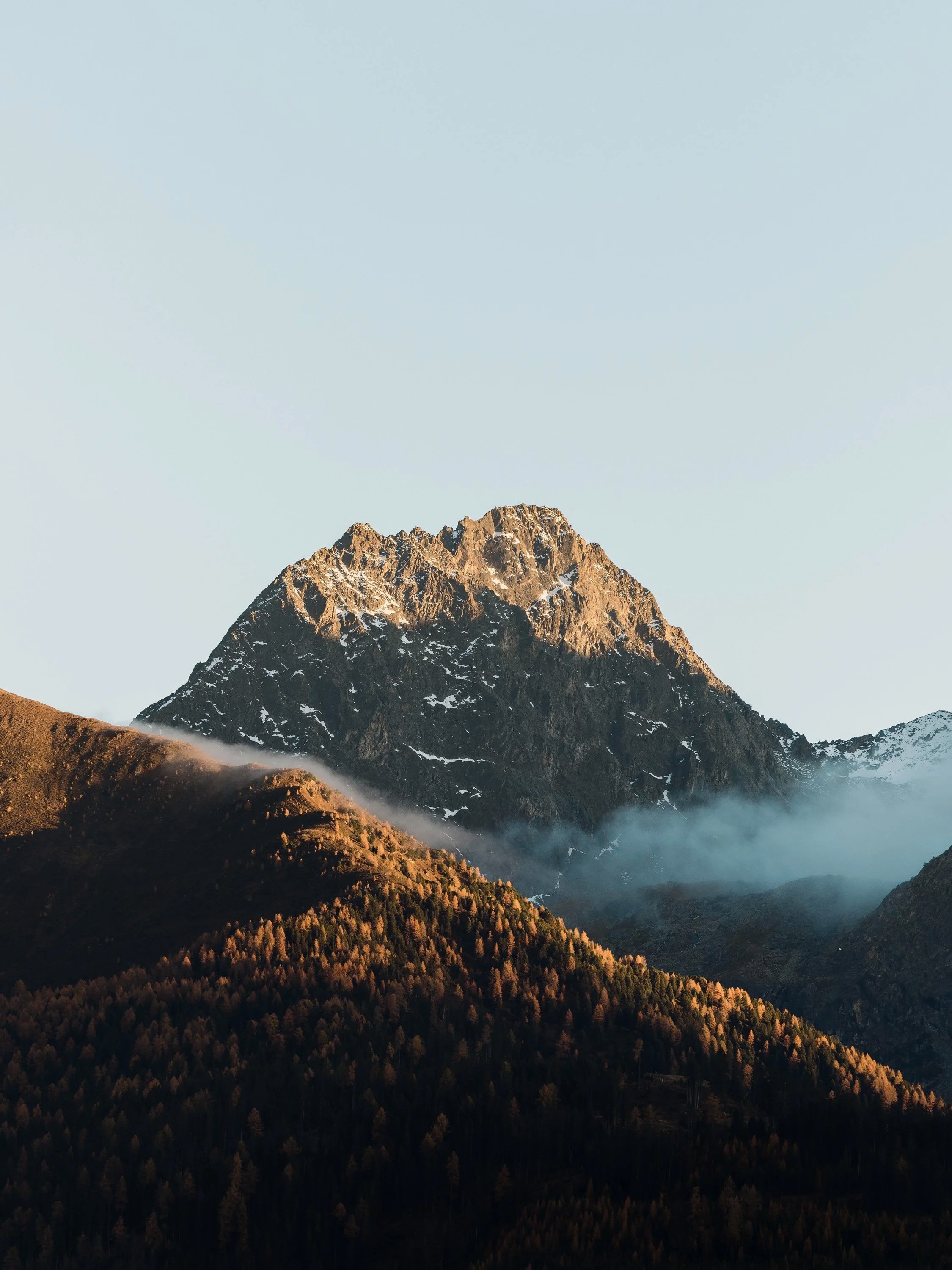 Berglandschaft mit steiler, felsige Bergspitze im Hintergrund und bewaldeten Hügel im Vordergrund, bei Sonnenaufgang oder -untergang, mit leichter Nebelbildung.