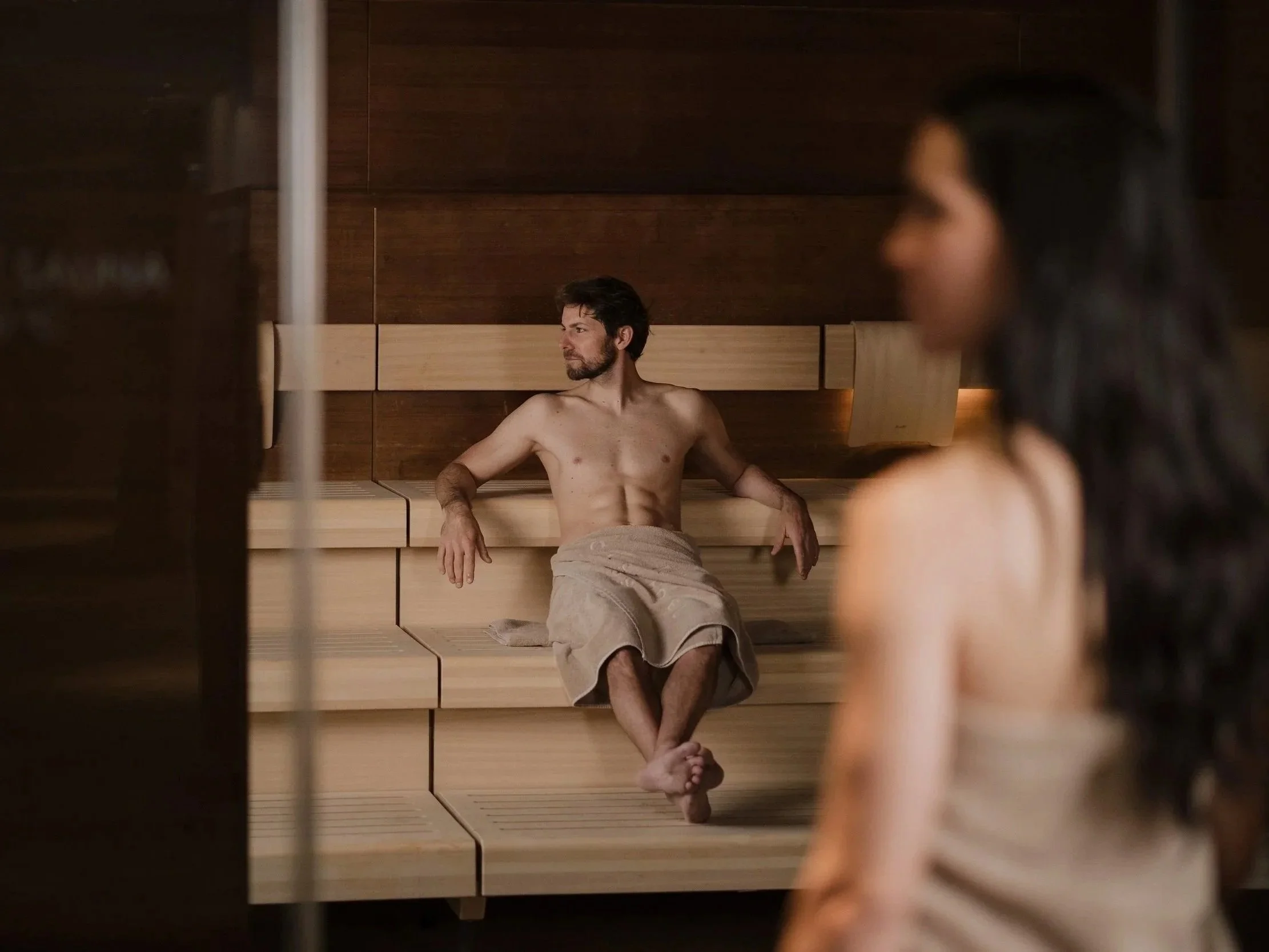 Ein Mann sitzt in einem Saunaraum, umgeben von Holz, mit einer Hand auf der Lehne und einer Tasse auf dem Boden. Im Vordergrund ist eine Frau mit dunklen Haaren, die nur teilweise sichtbar ist, ebenfalls in der Sauna.