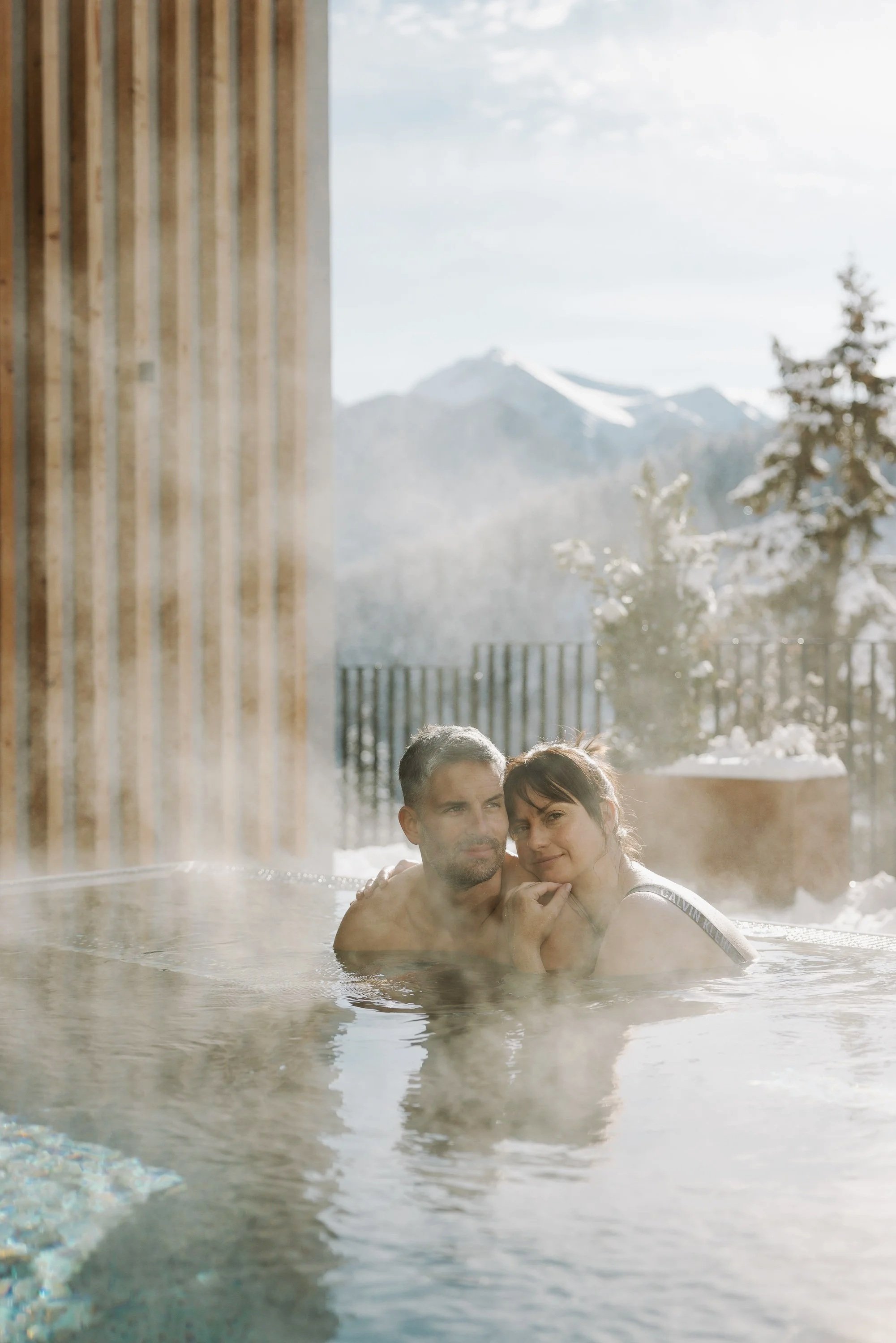 Ein Paar entspannt im warmen Außenwhirlpool inmitten von Schnee, mit Blick auf schneebedeckte Berge und Tannen, umgeben von Natur.
