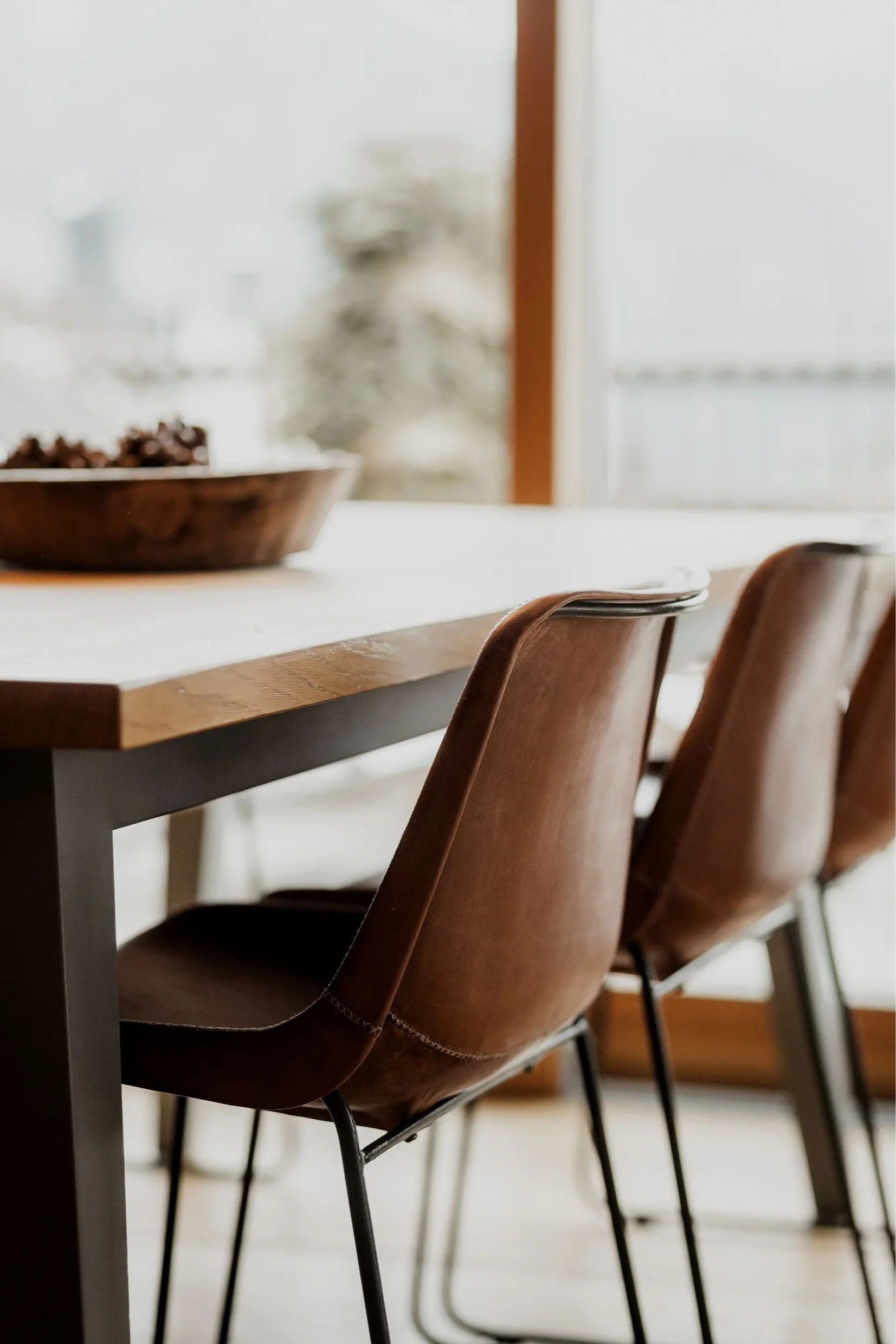 Ein Esszimmer mit einem Holztisch, braunen Stühlen und einem großen Fenster im Hintergrund. Auf dem Tisch befindet sich eine Schale mit Trauben.