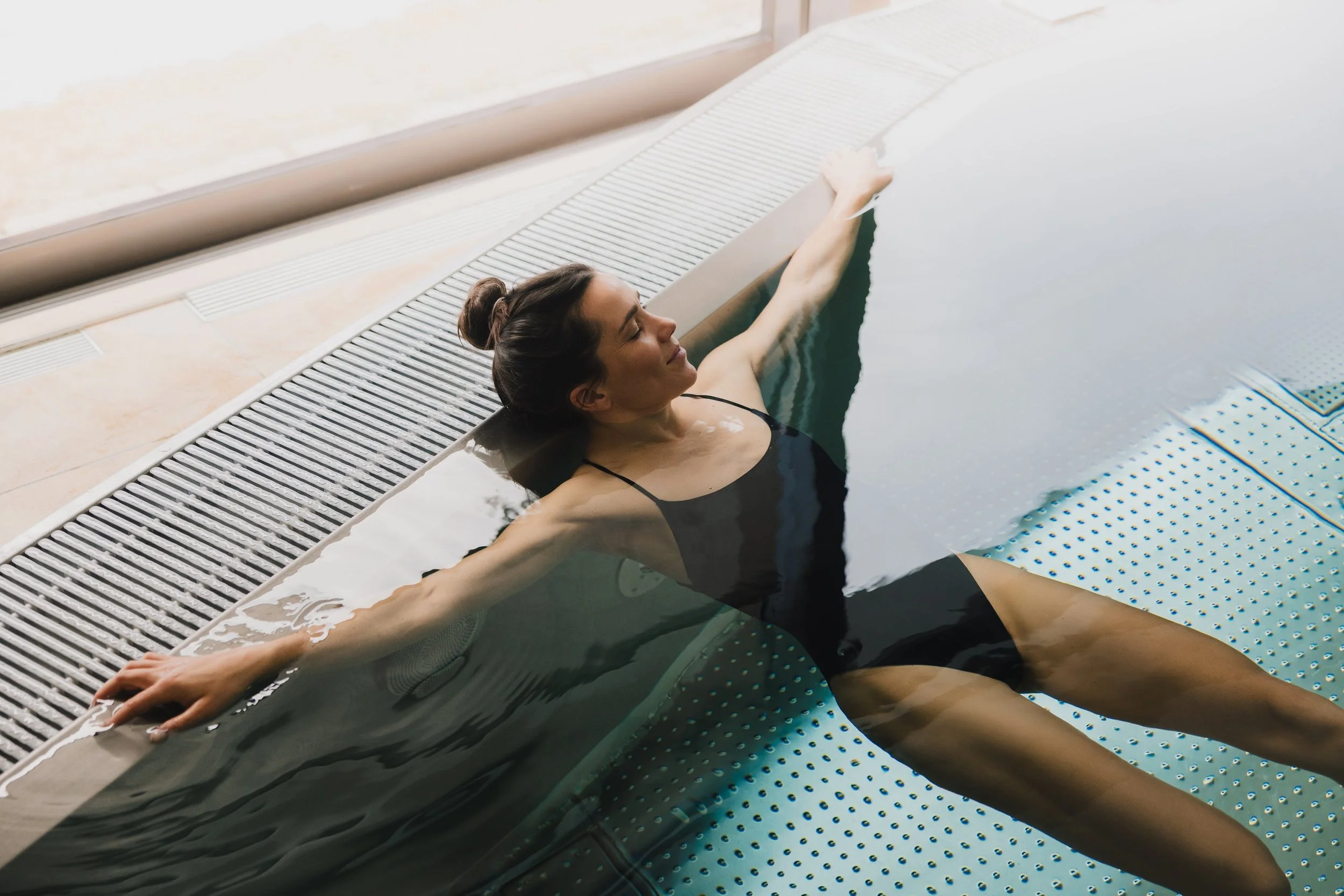 Frau entspannt sich im Spa-Pool, sitzt mit ausgestreckten Armen im Wasser, trägt einen schwarzen Badeanzug, mit dunkelbraunen Haaren im Dutt, in einem modernen Wellnessbereich.
