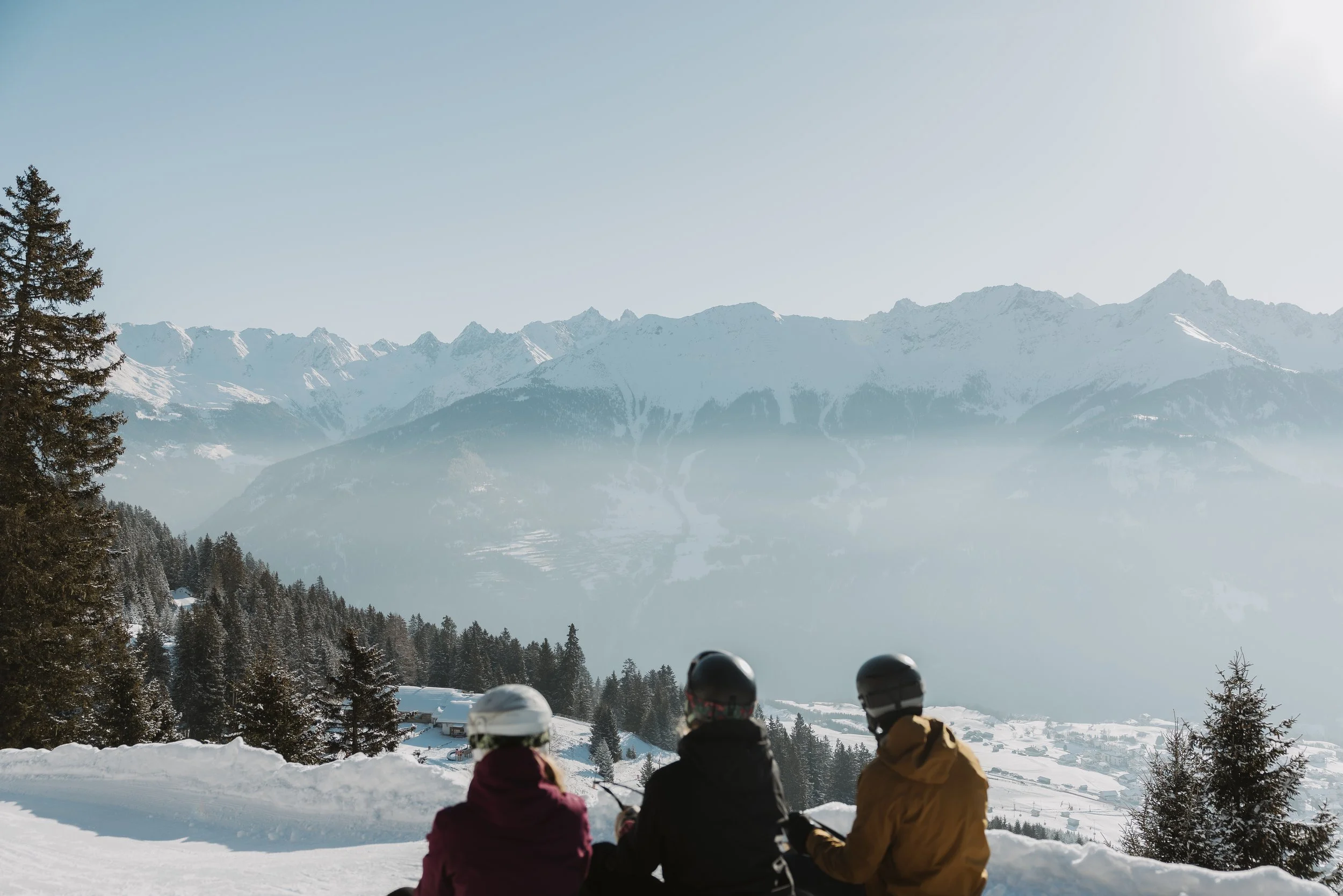 Drei Personen in Winterkleidung und Helmen sitzen im Schneidersitz auf einem verschneiten Hang und schauen auf eine verschneite Berglandschaft mit hohen Gipfeln und Nebel in der Morgendämmerung.