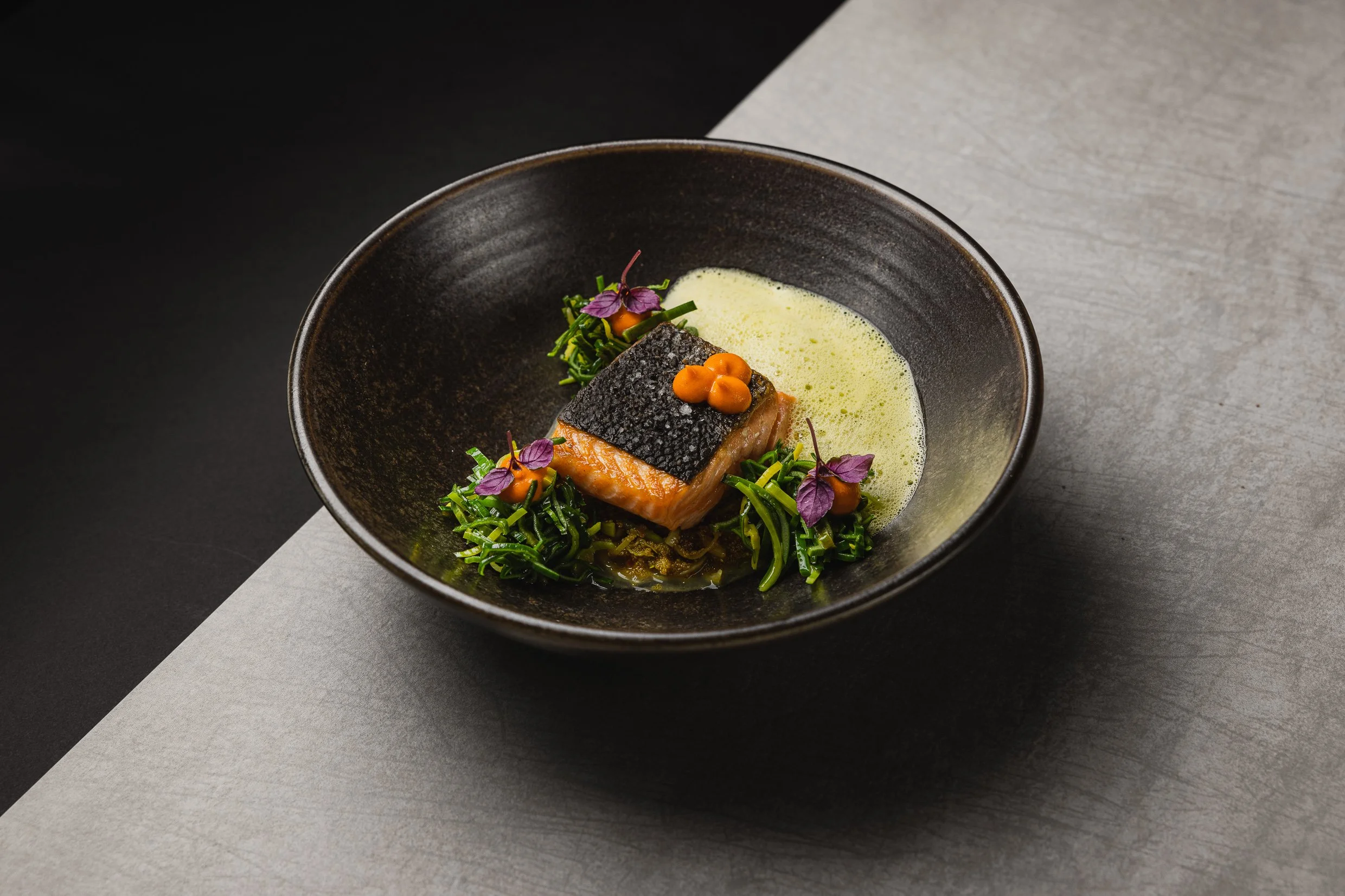 Gegrilltes Lachsfilet mit schwarzer Haut, serviert auf einem schwarzen Teller mit grünen Gemüse und einer cremigen, gelben Sosse, garniert mit kleinen orangefarbenen Tupfen und lila Blättern.