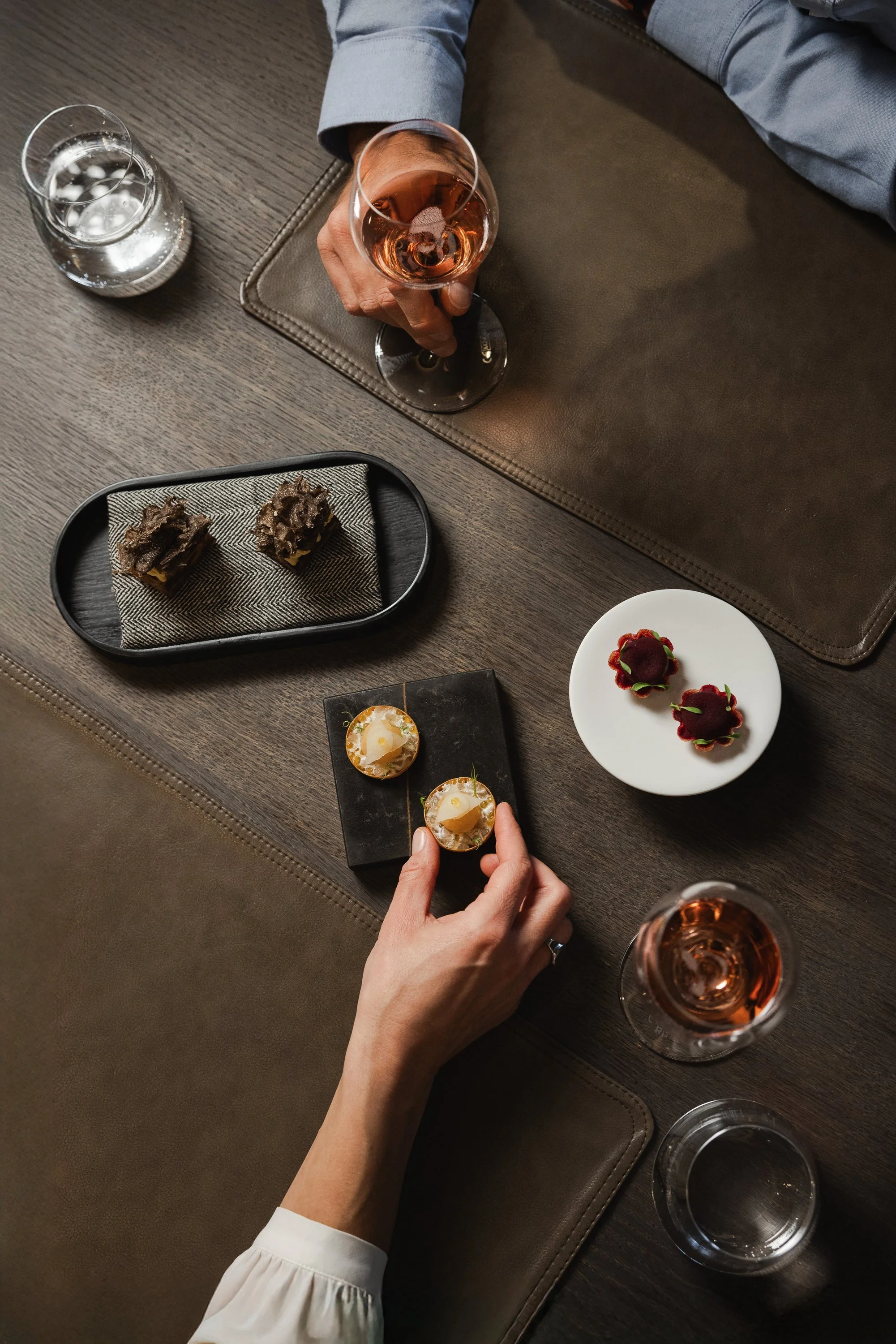 Tisch mit Desserts und Getränken, eine Hand hält ein Glas Roséwein, die andere hält ein kleines Dessert. Es gibt zwei Stücke Dessert auf einem kleinen schwarzen Teller, zwei weitere auf einem weißen Teller, Wasser und eine zweite Glaskaraffe.