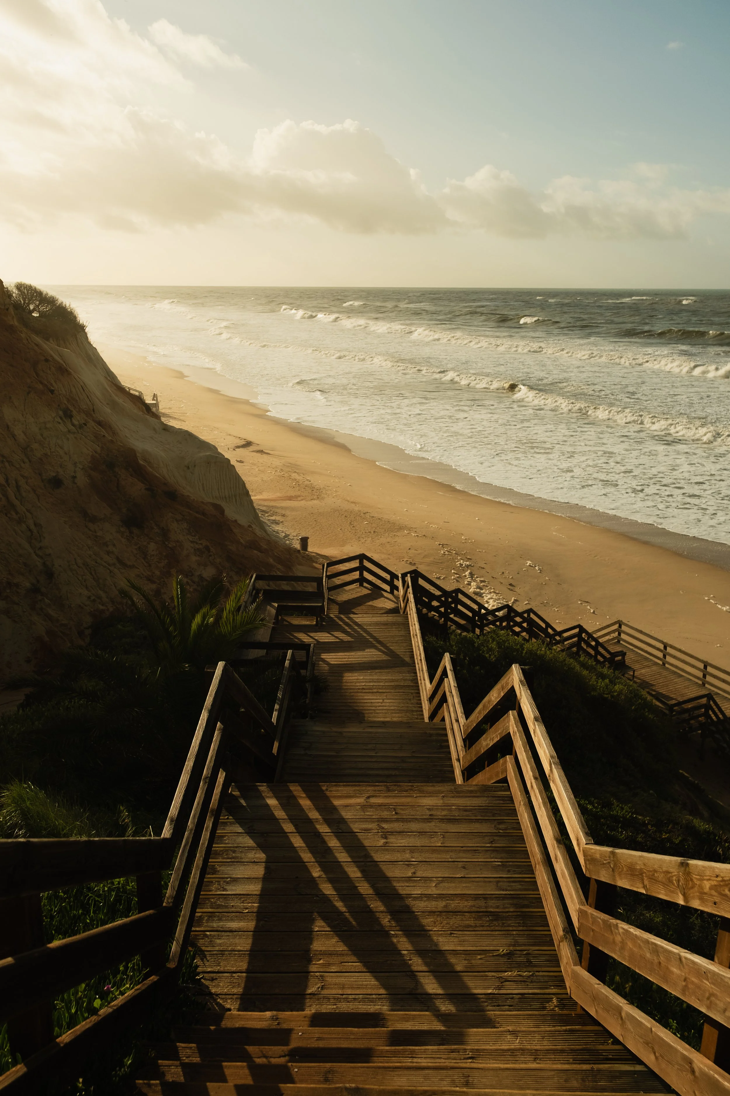 Holztreppe führt vom Strand zu einer Klippe, Blick auf das Meer bei Sonnenuntergang, mit Wolken am Himmel.