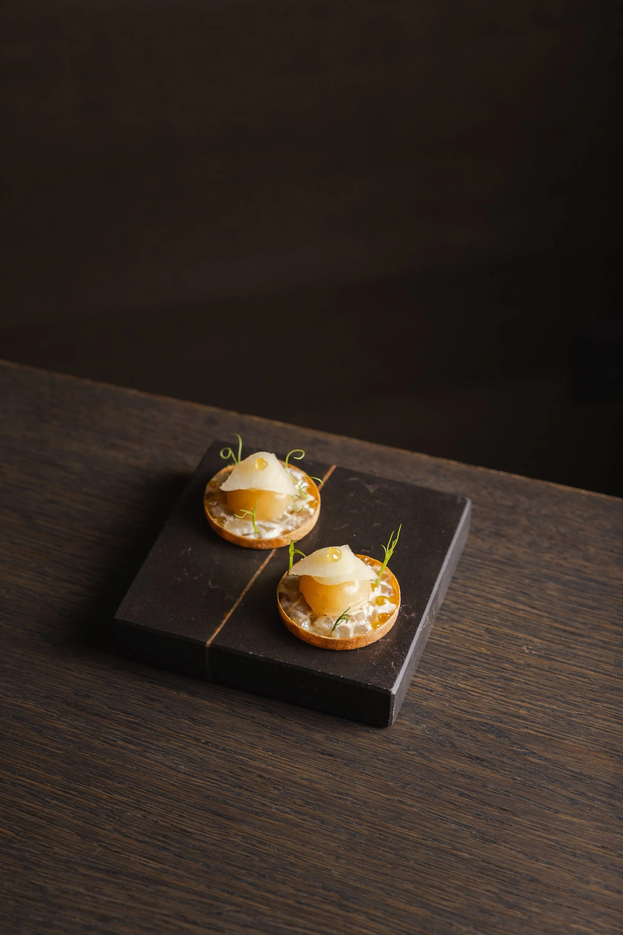 Zwei kleine Tartelette mit hellem Creme, einer Käse- oder Fruchtmischung, garniert mit kleinen grünen Kräutern, auf einer schwarzen Steinplatte auf einem dunklen Holztablett.