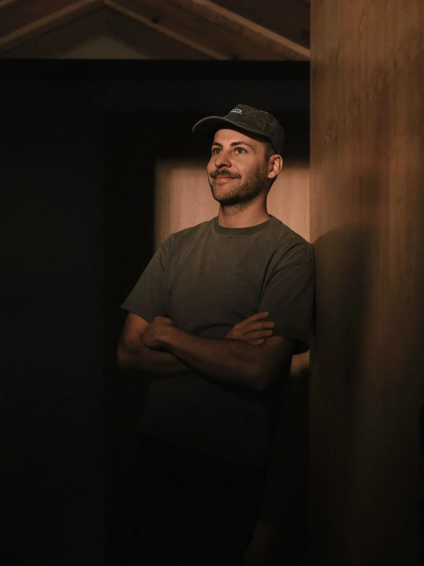 Ein Mann lehnt an einer Holzwand in einem dunklen Raum, trägt ein graues T-Shirt, eine schwarze Cap und sieht nach rechts mit einem leichten Lächeln.