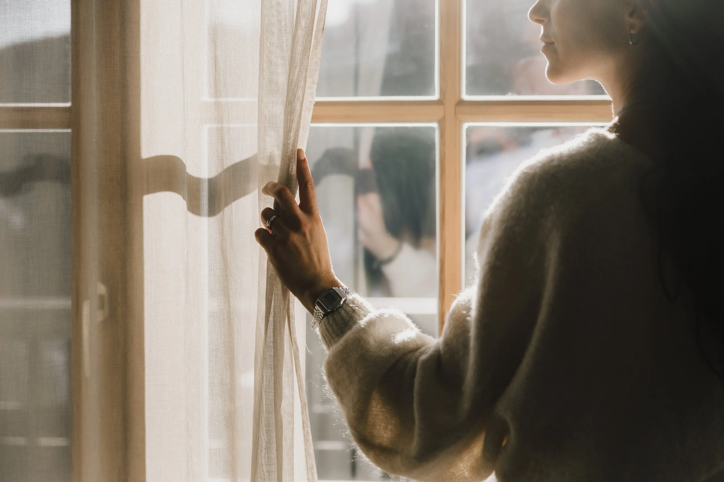 Frau zieht einen Vorhang in einem sonnendurchfluteten Raum beiseite, während sie nach draußen schaut.