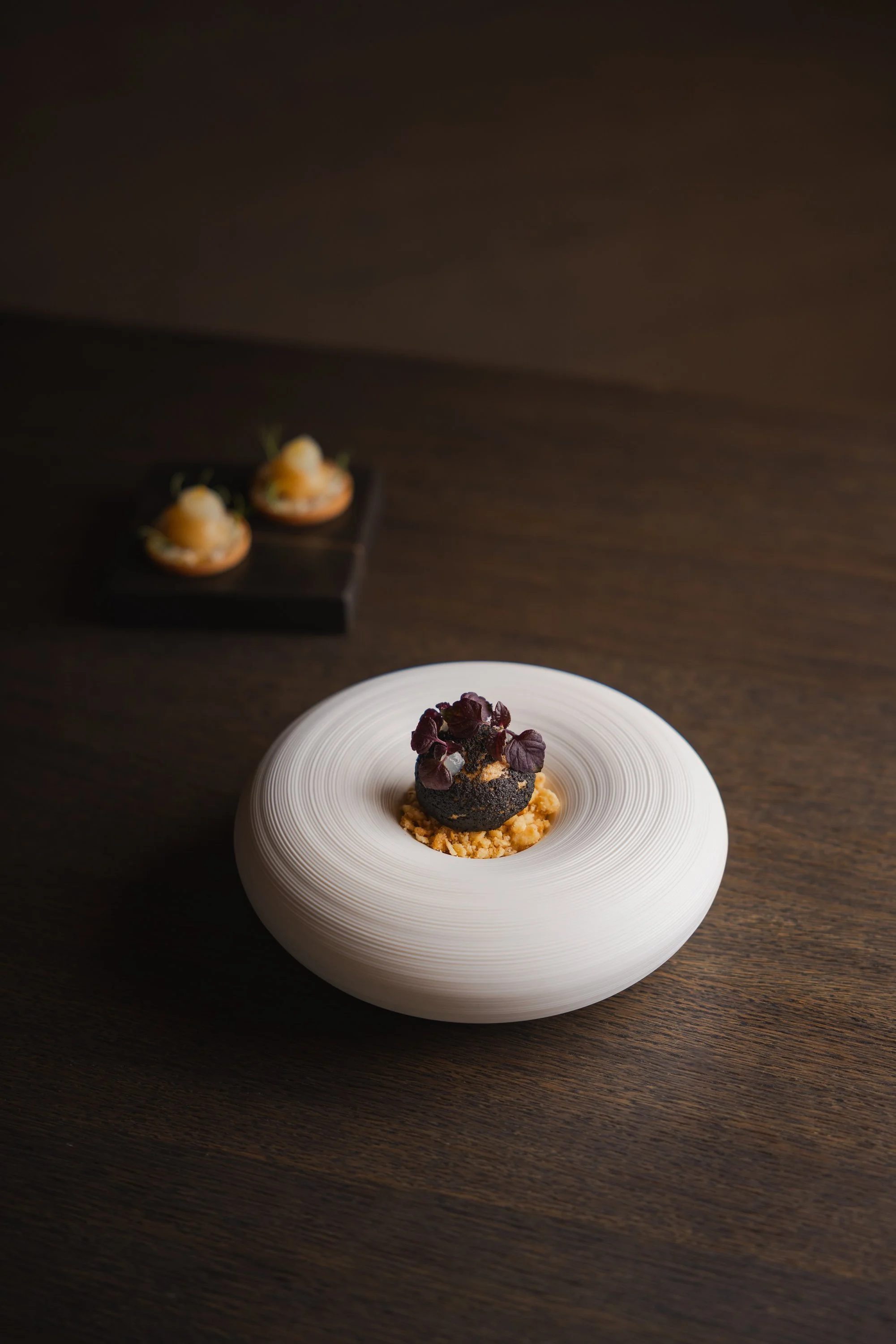 Elegantes Dessert auf einer weißen, runden keramischen Schale mit spiralförmigem Muster, dekoriert mit schwarzen und lila Blättern, auf einem dunklen Holzuntergrund. Im Hintergrund sind zwei kleine Häppchen auf einem schwarzen Brett. 