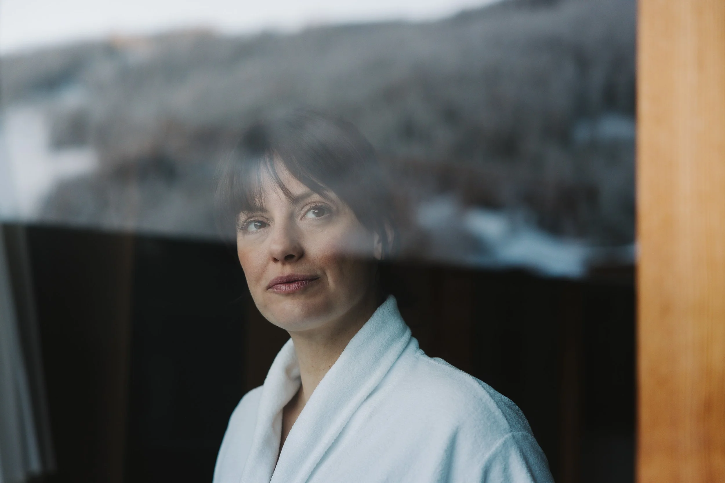 Junge Frau schaut nachdenklich durch ein Fenster, trägt einen weißen Bademantel.