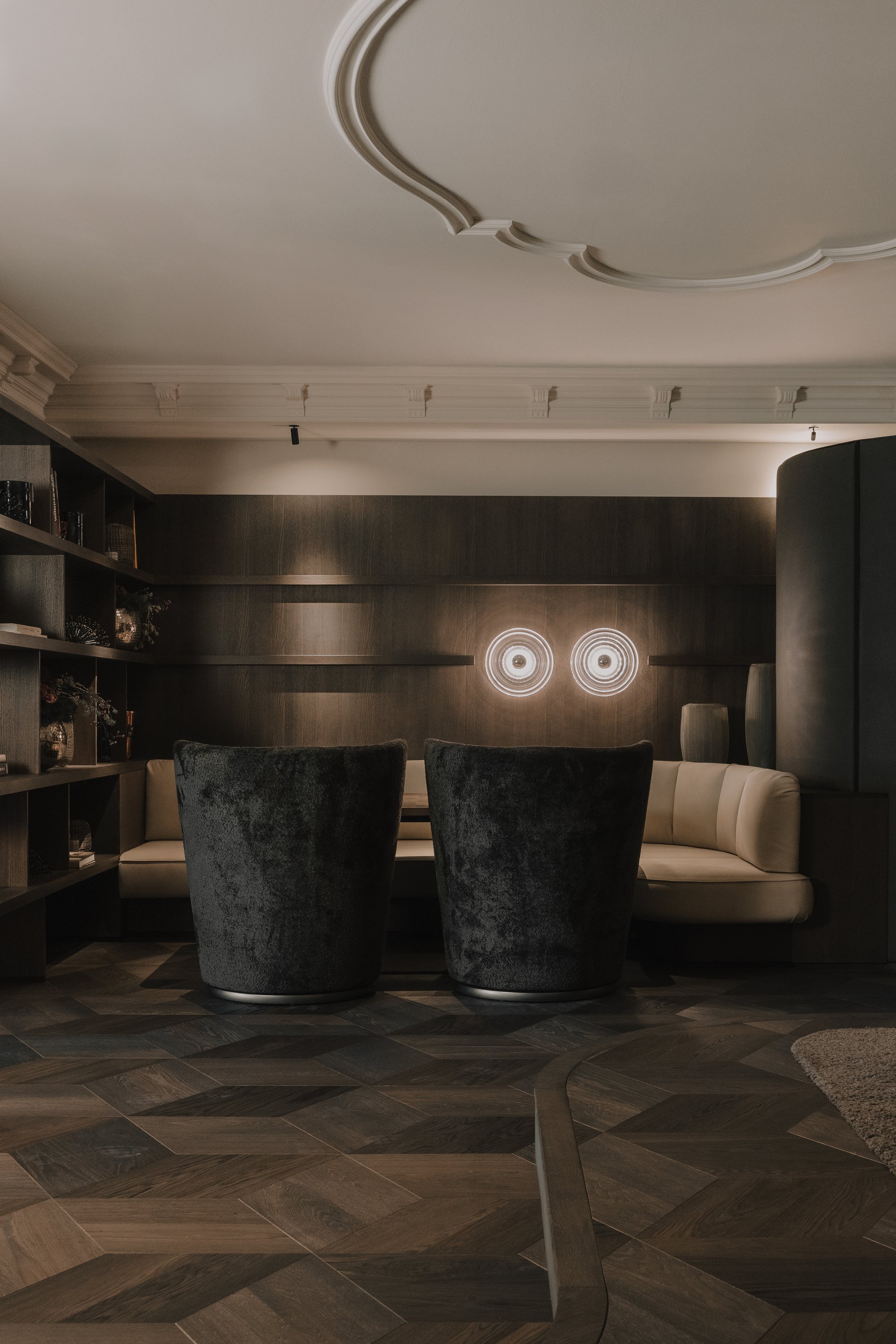 Elegantes Wohnzimmer mit dunklen Wänden, schwarzen hohen Sesseln, einem cremefarbenen Sitzbereich, Bücherregale, modernen Wandlampen und dunklem Holzboden.