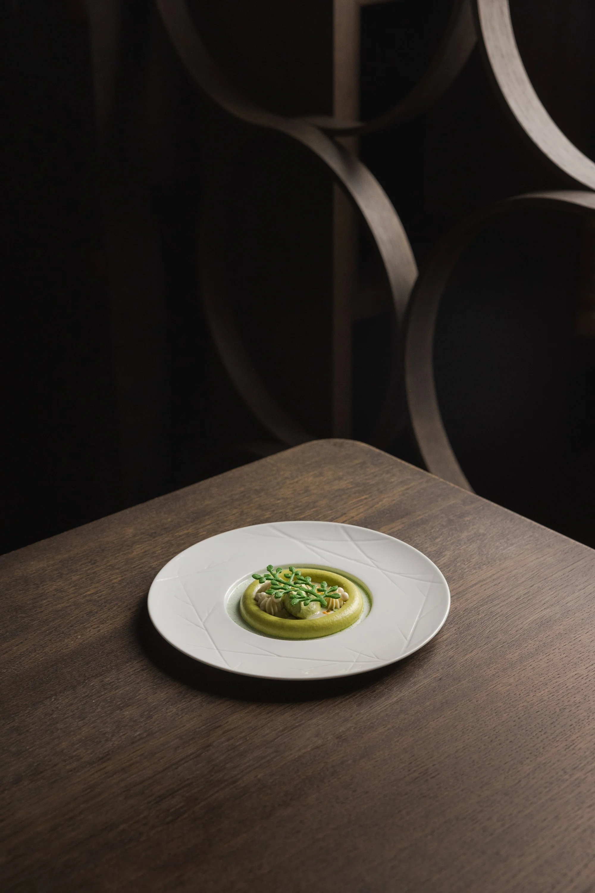 Ein elegantes Gericht mit grünen Soßen und Kräutern auf einem weißen Teller auf einem Holztisch, dunkler Hintergrund.