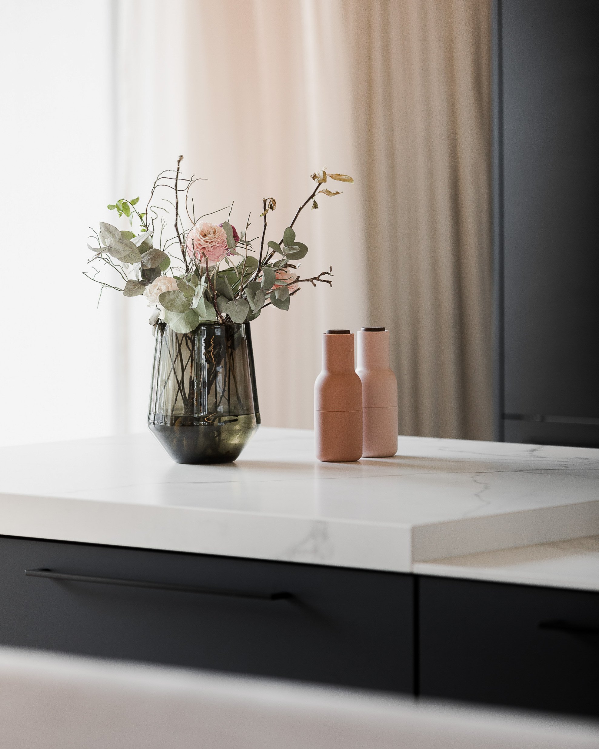 Blumen in einer schwarzen Vase neben zwei rosa Flaschen auf einer weißen Tischplatte in einer modernen Küche.