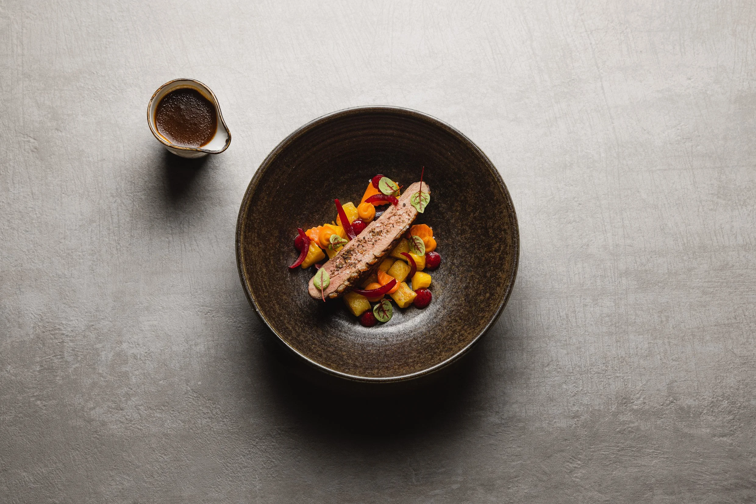 Ein Teller mit einem Stück Fleisch oder Fisch auf einem Gemüsebett, serviert mit einer dunklen Soße, auf einem grauen Teller mit einer kleinen Sauciere daneben, alles auf einer hellen, strukturierten Oberfläche.