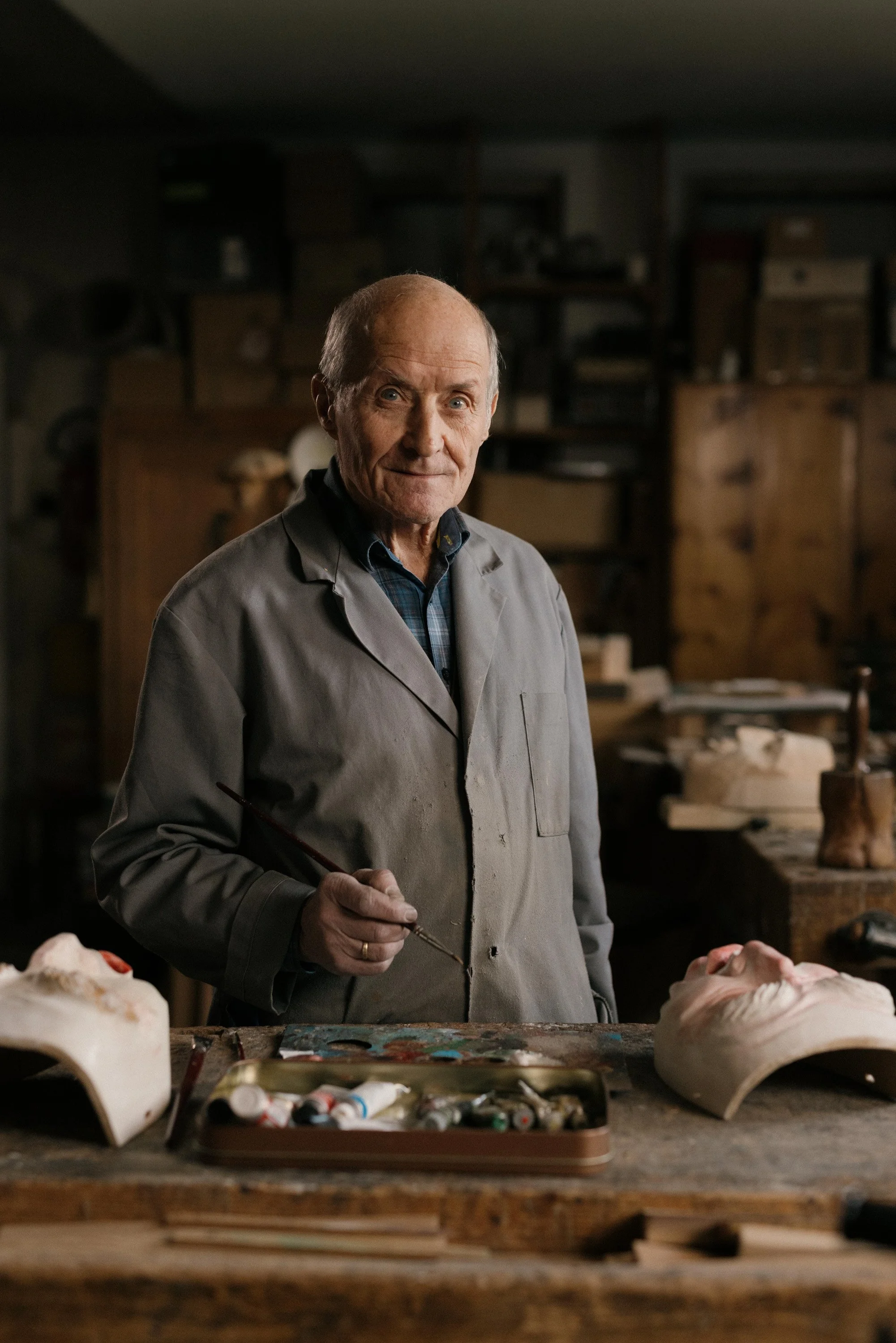 Ein älterer Mann in einem Atelier mit Masken und Malutensilien, der einen Pinsel hält.