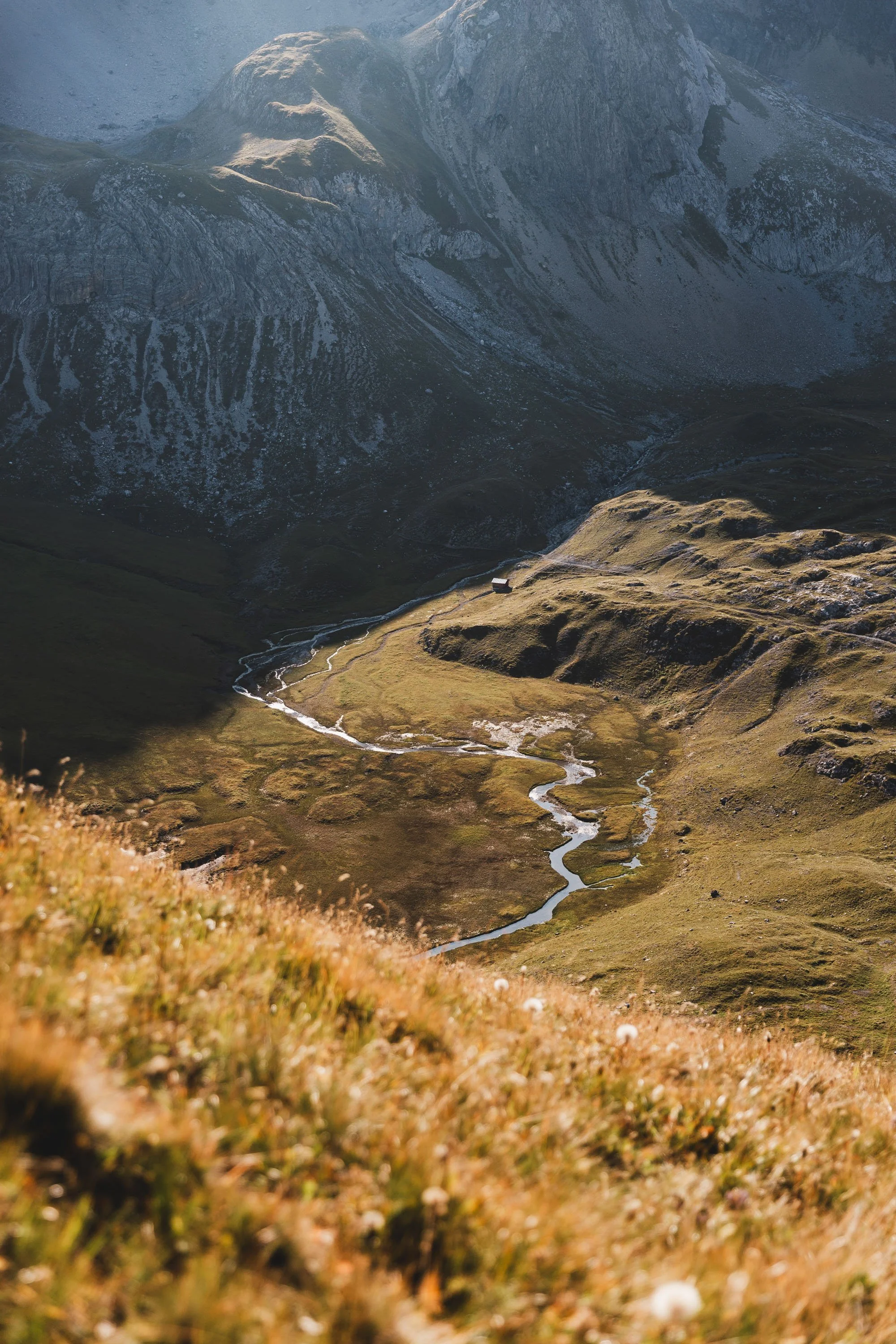 Ein Bergpanorama mit einer kleinen durch den Fluss verlaufenden Wiese im Tal, umgeben von hohen Bergen mit felsigen und grasbedeckten Flächen